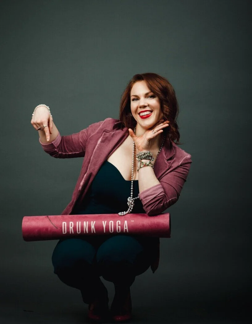 Eli Walker, a woman with red hair, wearing a pink blazer and black top, sitting with a rolled yoga mat labeled 'Drunk Yoga.' She is smiling, with her left hand near her chin, and her right arm flexed, showing her bicep.