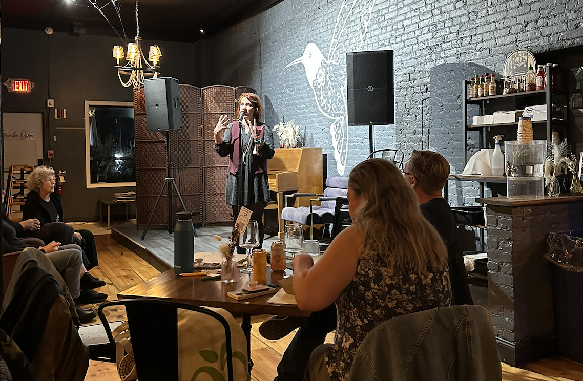 A woman is performing stand-up comedy on stage, holding a microphone in a cozy, dimly lit venue with an audience seated at tables.