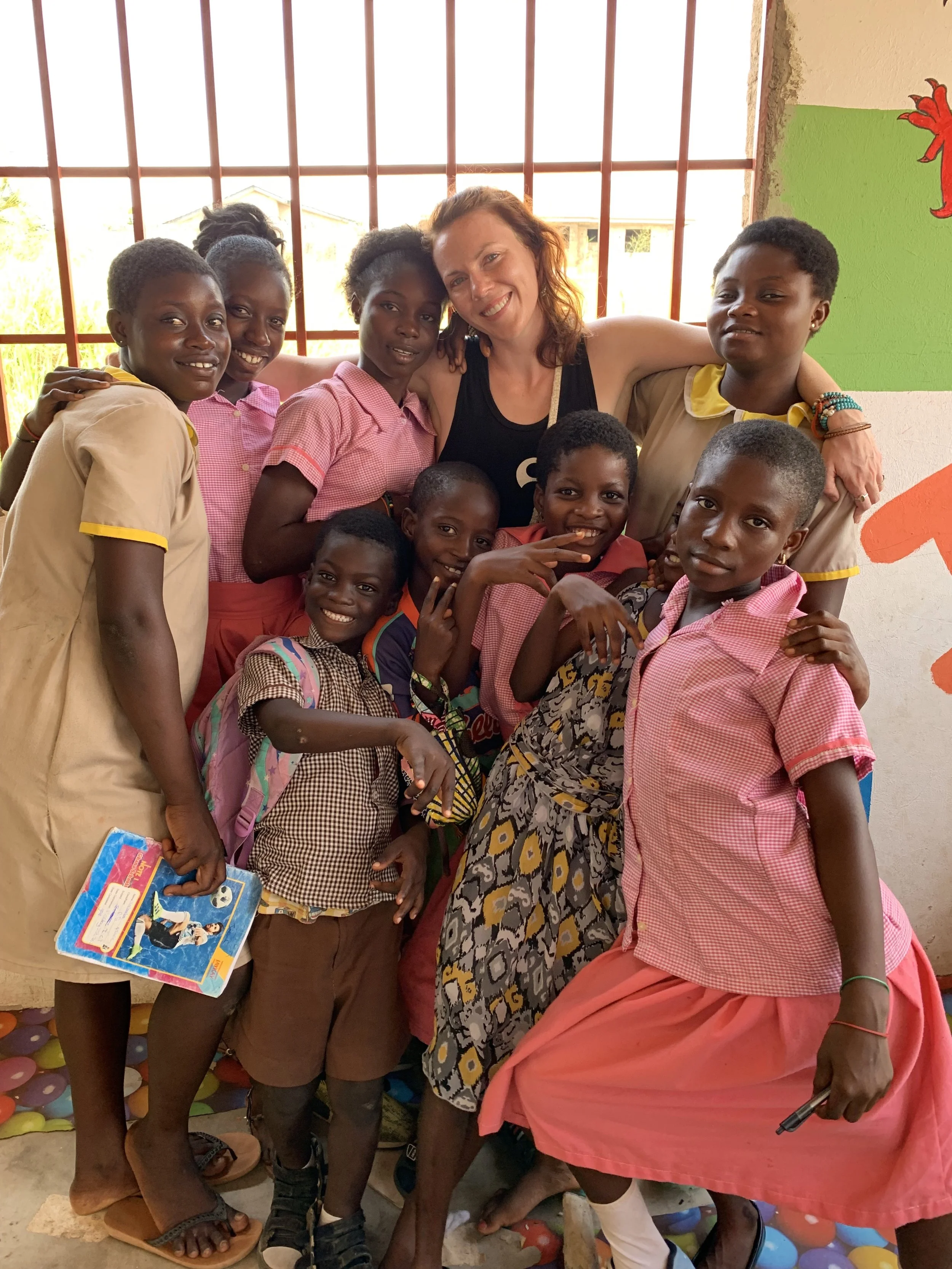 A group of children and two women smiling and posing together indoors with a brightly lit window in the background.
