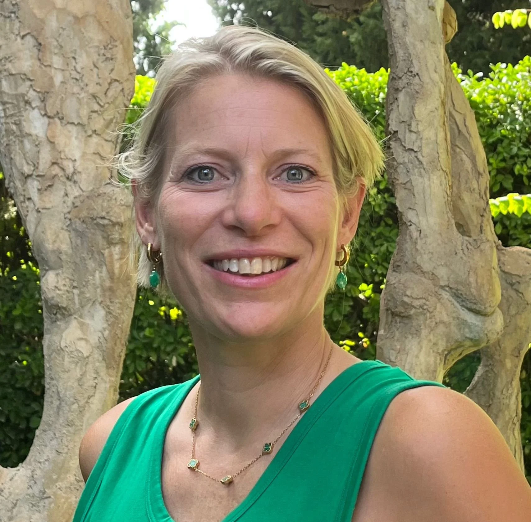 A woman with short blonde hair wearing a green sleeveless top, necklace, and earrings, smiling outdoors in front of a tree and greenery.