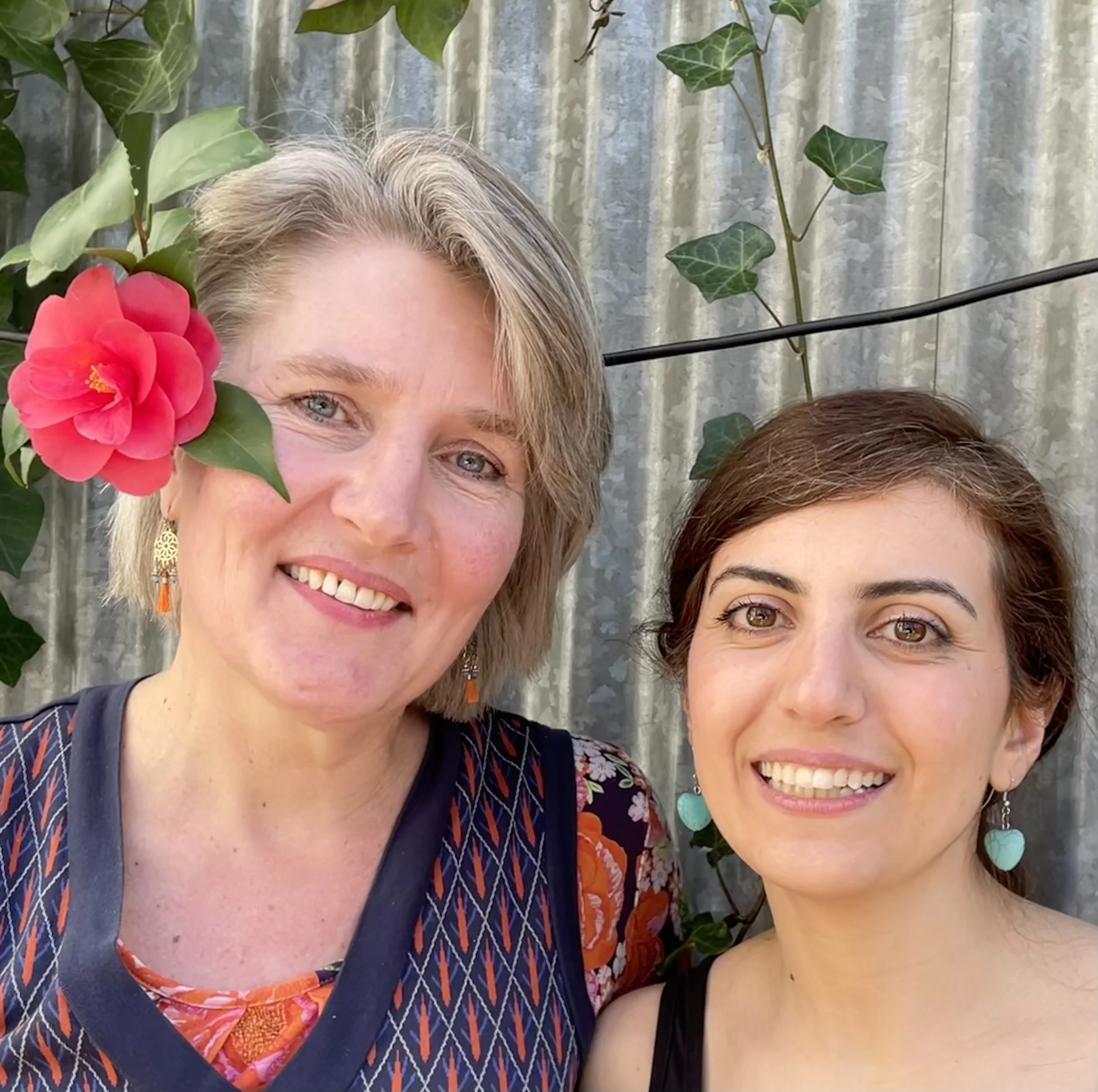 Two women smiling outdoors, with greenery and a metal fence in the background. One woman has short blonde hair, wears earrings, and a floral top. The other has short dark hair, earrings with heart-shaped pendants, and a sleeveless black top. A pink flower and green leaves are visible near the blonde woman.