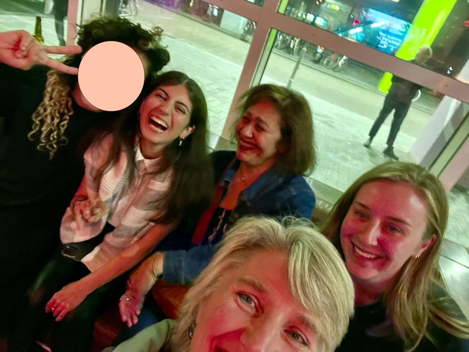 Group of five women taking a selfie together, all smiling and enjoying a night out, in a room with large windows and a view of a street outside.