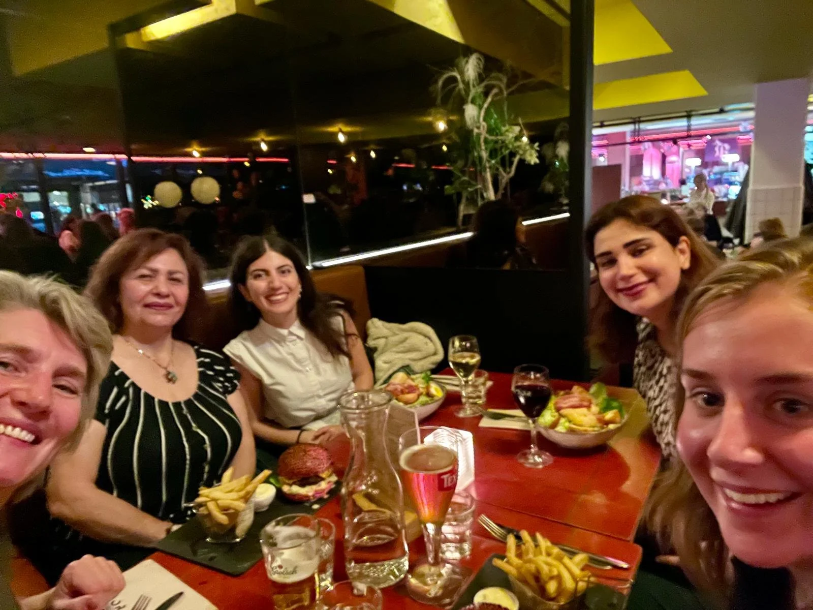 A group of six people, five women and one man, sitting at a table in a restaurant, smiling at the camera. The table has plates of food, glasses of wine, and a pitcher of water. The background shows a lively restaurant setting with other patrons and colorful lighting.