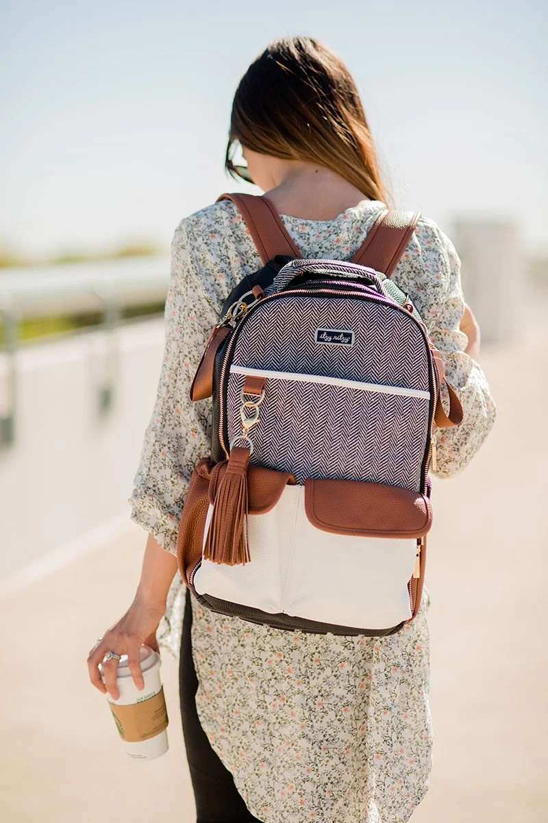 A woman walking outdoors on a sunny day, holding a coffee cup, wearing a floral dress and carrying a stylish backpack.