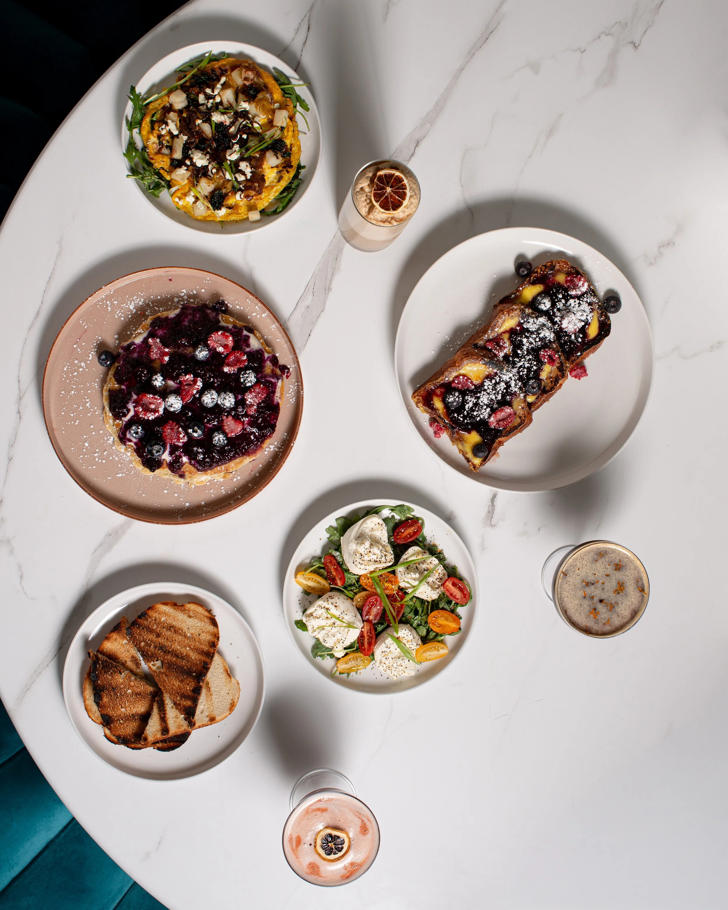 Top-down view of a white marble table with various dishes including a vegetable omelette, fruit tarts, a berry pastry, cheese and tomato salad, slices of toast, two glasses of beverages, and two bowls of drinks.