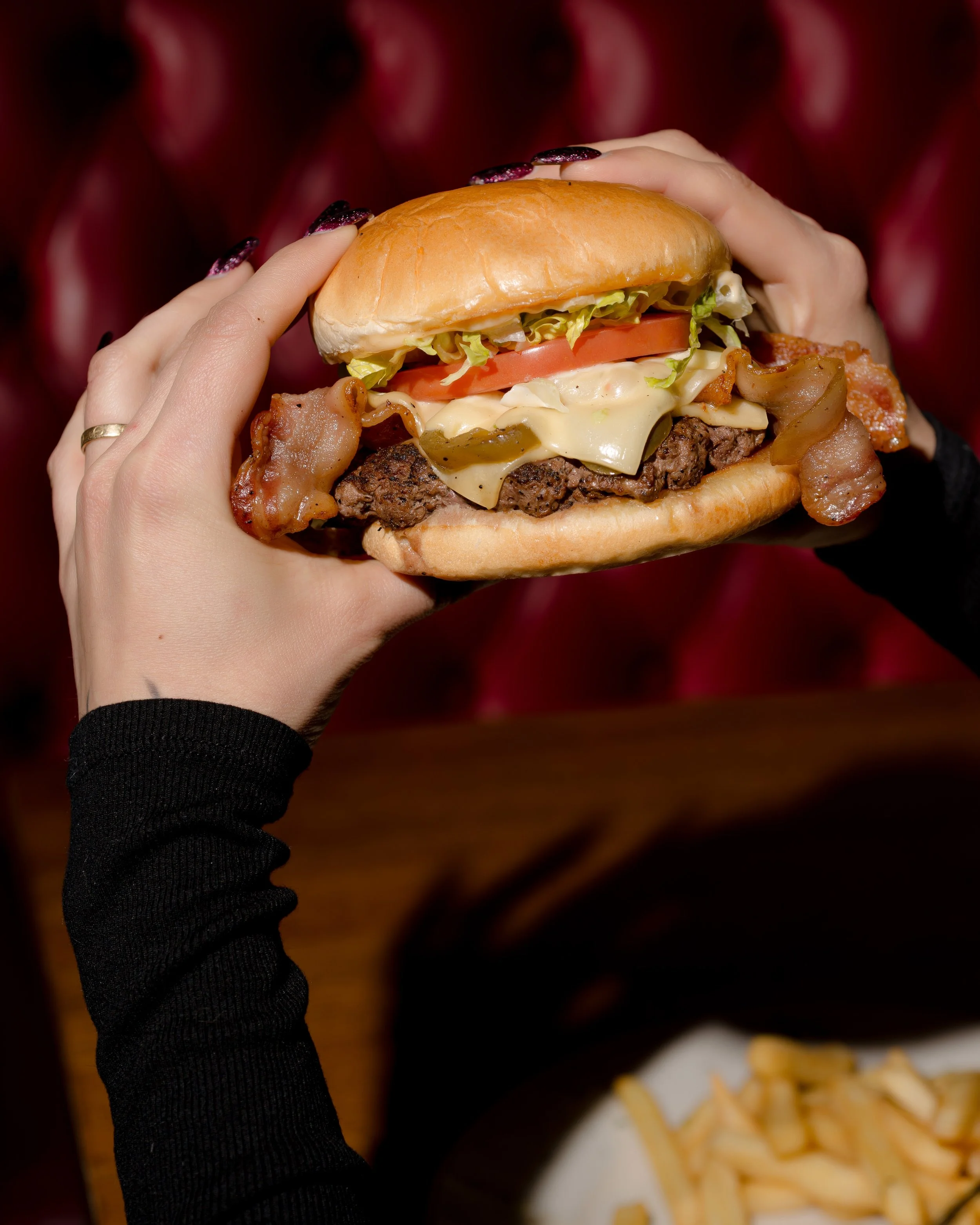 Person holding a bacon cheeseburger with lettuce, tomato, and cheese, in a restaurant setting.