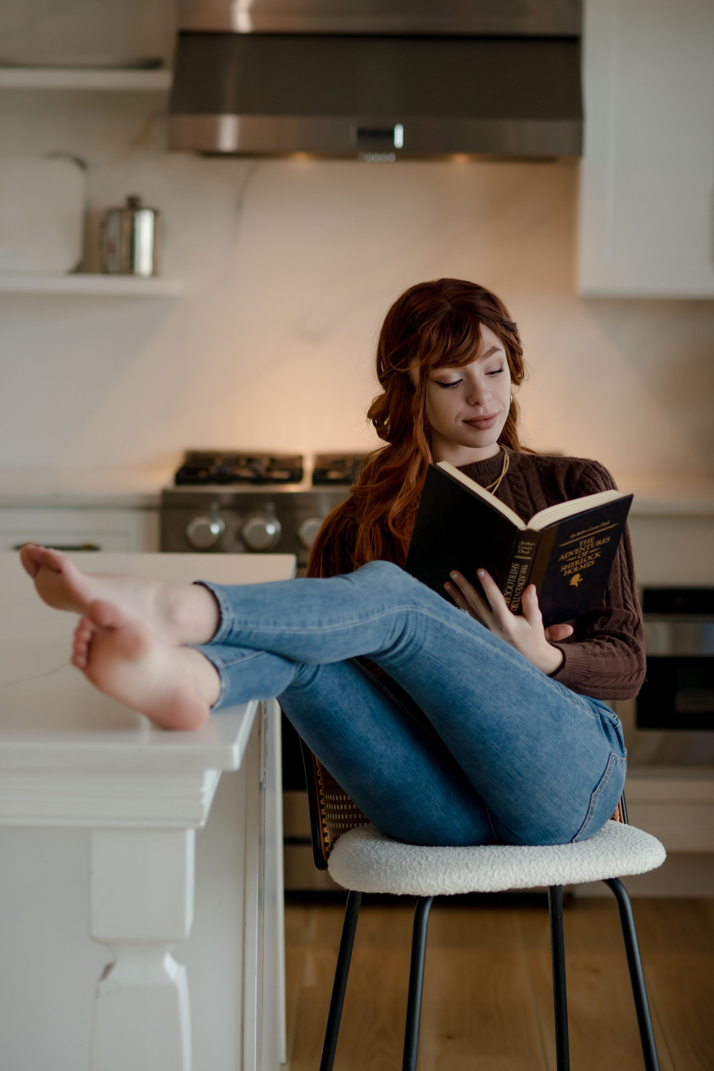 A young woman with red hair and casual clothing sitting on a chair with her legs resting on a kitchen counter, reading a book titled "The Adventures of Sherlock Holmes" in a kitchen with a stove and appliances.