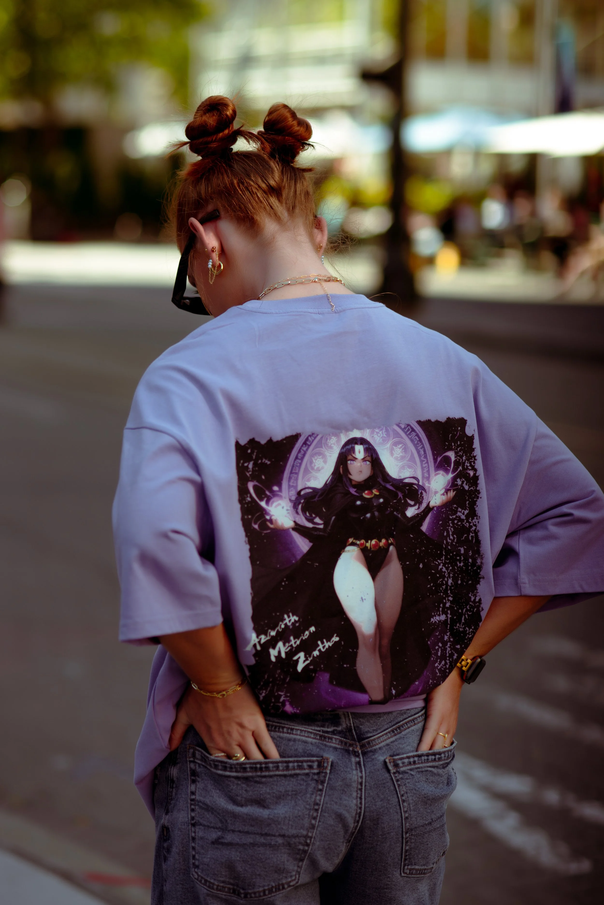 A woman with red hair styled in two buns is standing on a street with her back to the camera. She is wearing a purple T-shirt featuring an animated character with long black hair and glasses, surrounded by a cosmic background. She has earrings, layer