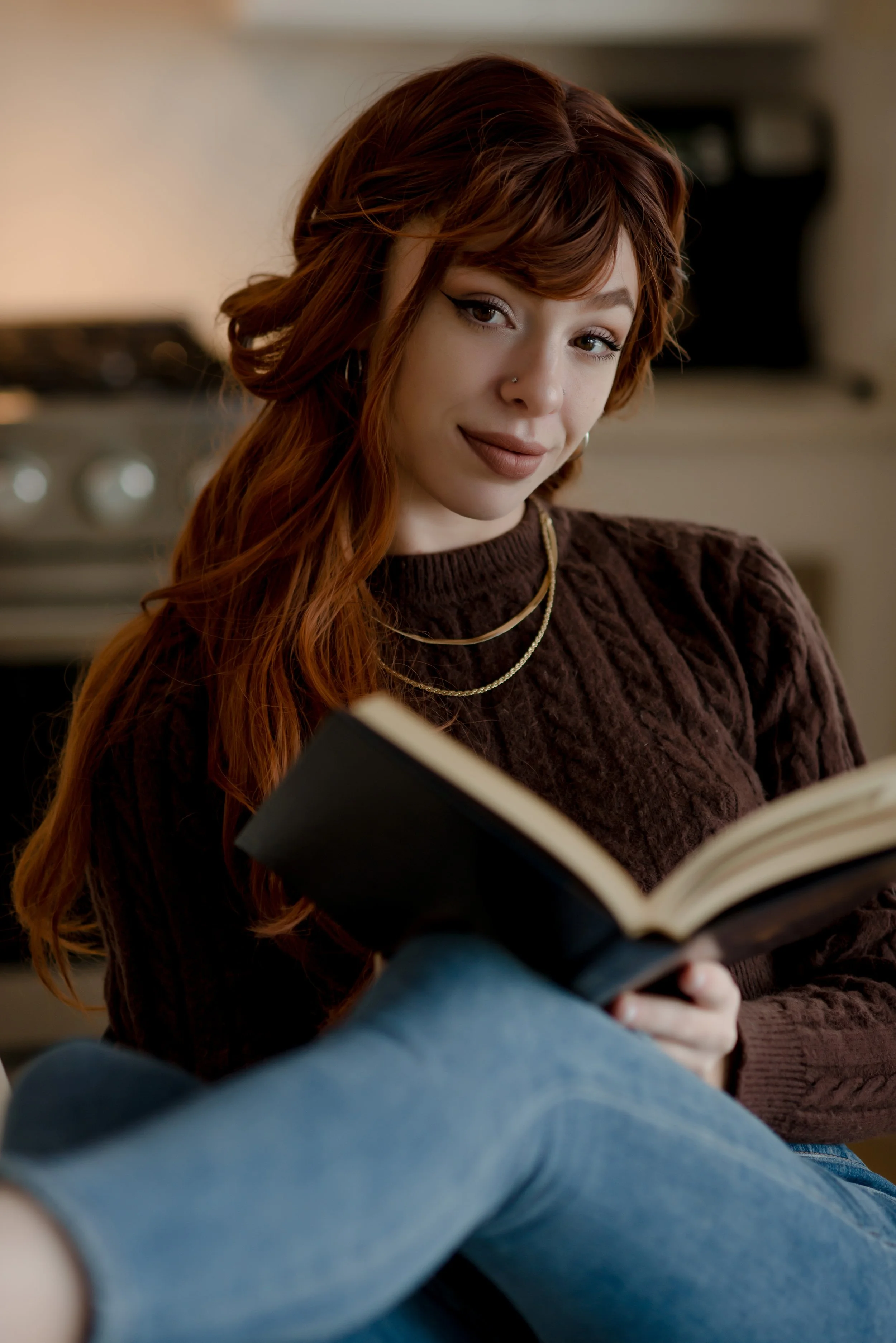 A young woman with red hair and a nose piercing, sitting cross-legged and reading a book in a cozy kitchen.