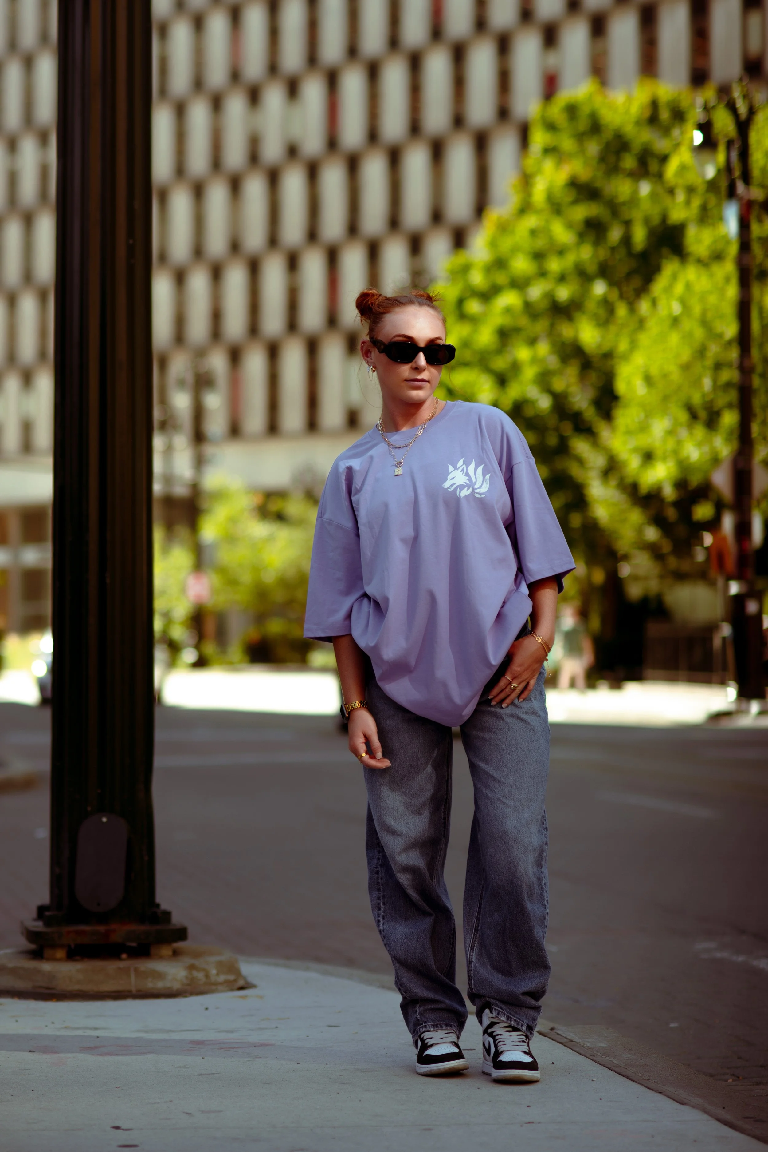 A woman with red hair tied in a bun, wearing dark sunglasses, a loose lavender t-shirt with a white cat logo, gray baggy jeans, and sneakers, stands on a city sidewalk with trees and buildings in the background.
