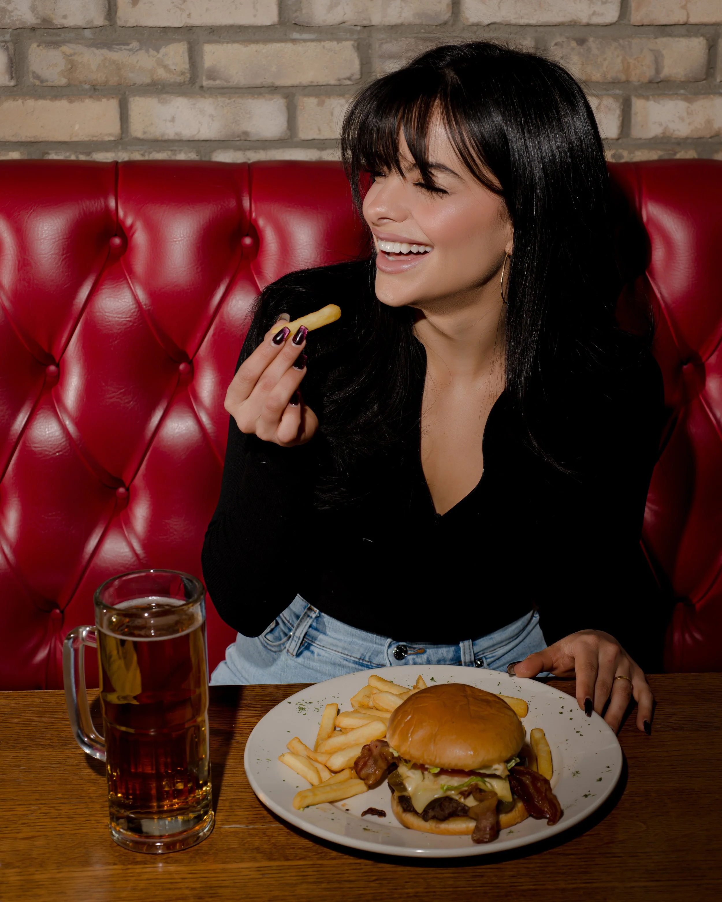 A woman with black hair and a black top smiling and holding a fry near her mouth at a restaurant. On the table in front of her is a plate with a cheeseburger, French fries, and a mug of beer.