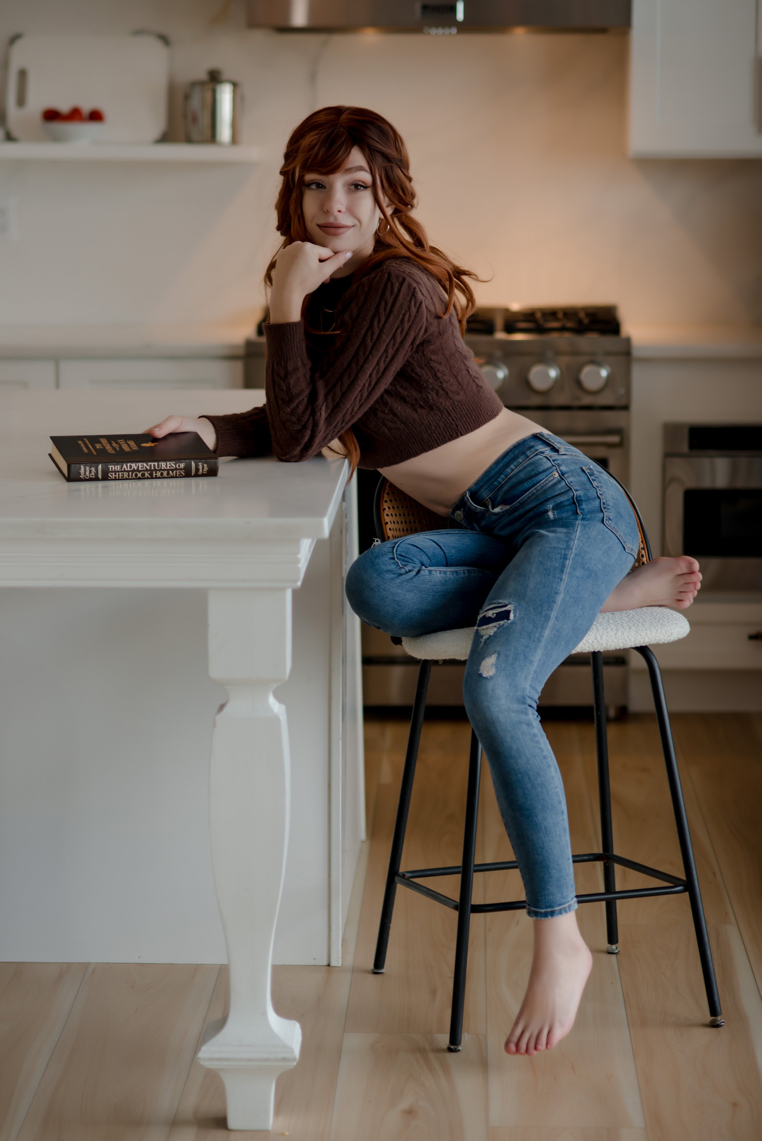 A woman with red hair sitting on a high stool at a kitchen island, wearing a cropped brown sweater and ripped jeans, with a book titled 'The Adventures of Sherlock Holmes' on the counter.