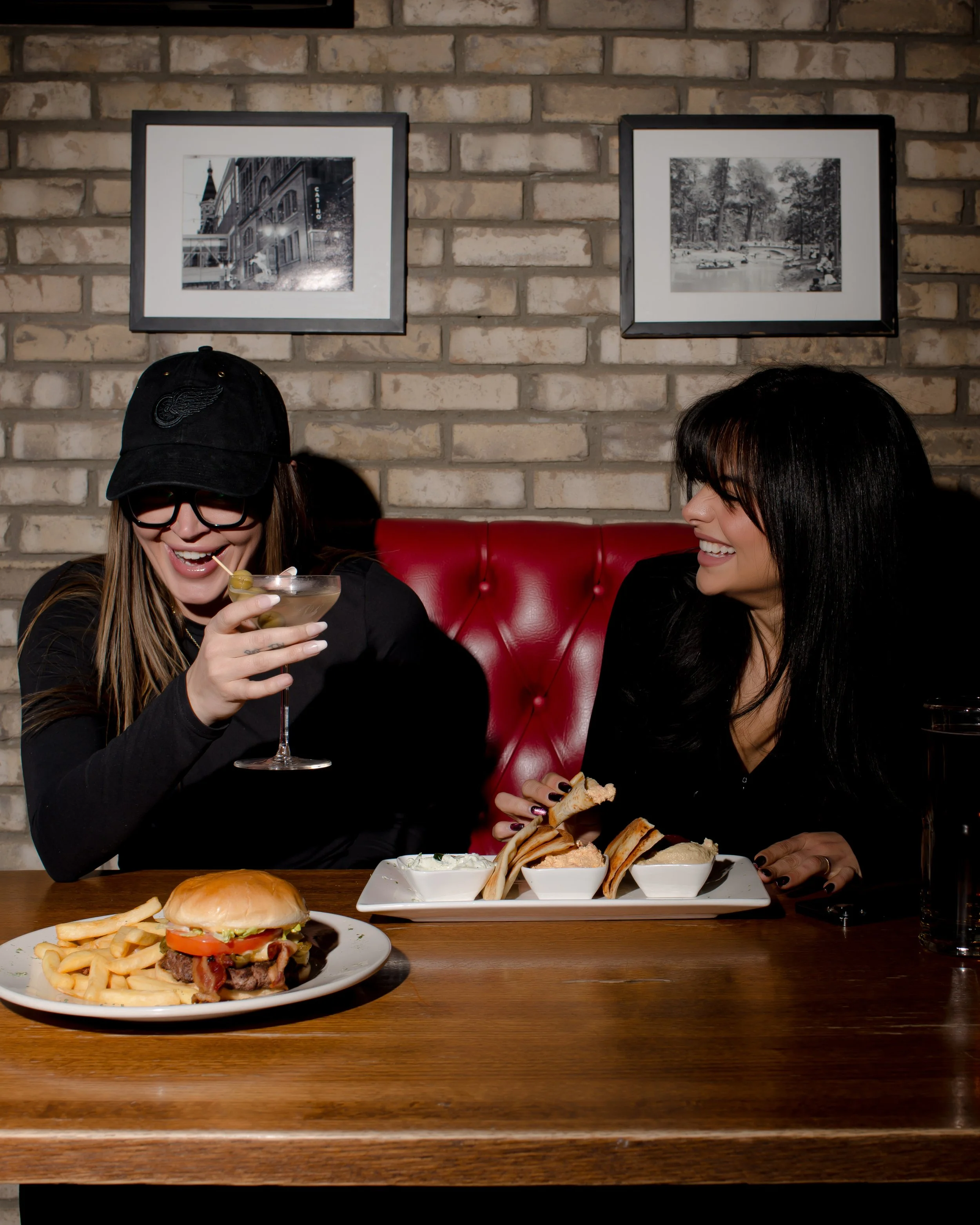 Two women enjoying food and drinks at a restaurant with a brick wall and framed black-and-white photos in the background.