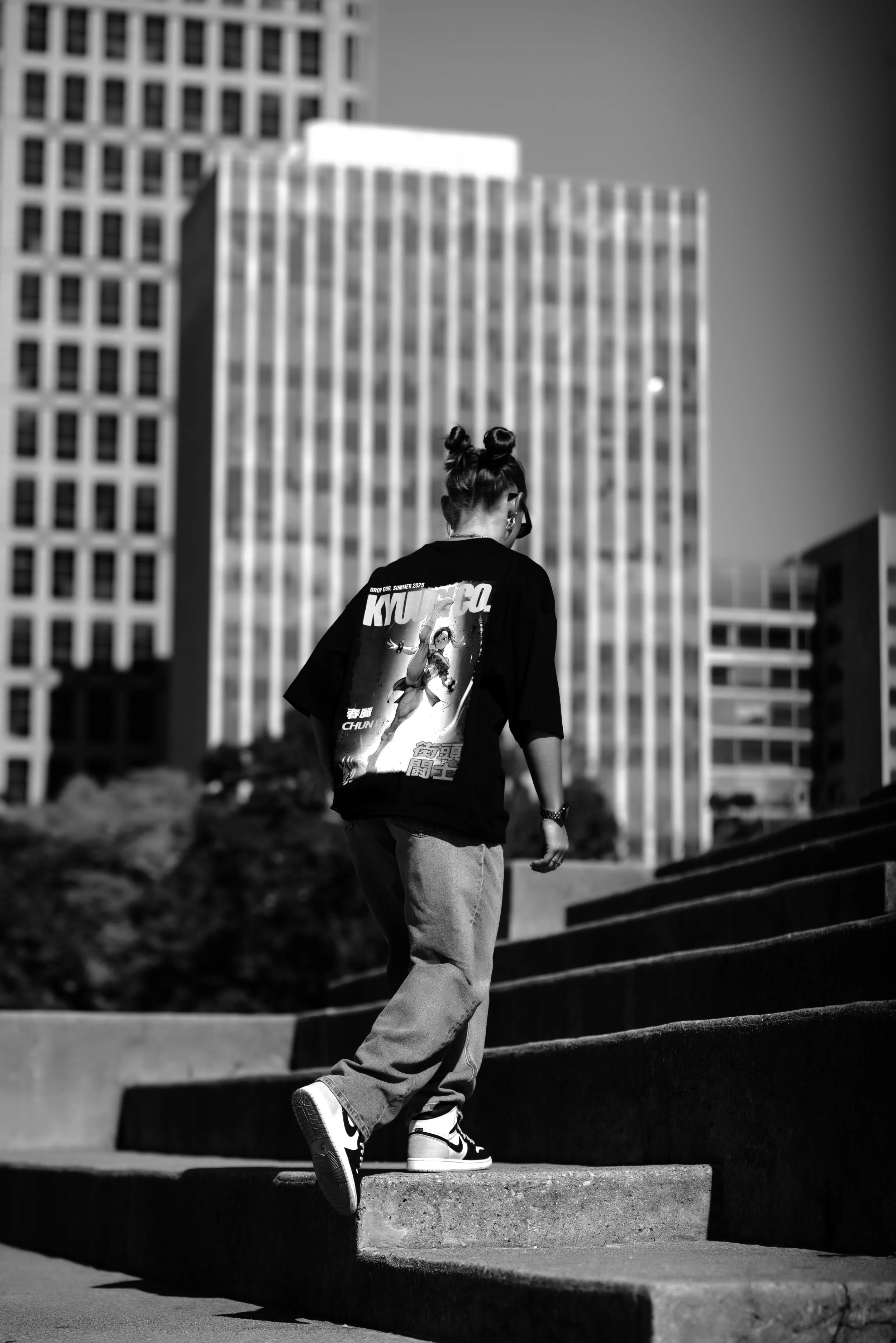 Person with two buns hairstyle, wearing a graphic T-shirt, jeans, and Nike sneakers, walking up concrete stairs in an urban area with tall buildings in the background.
