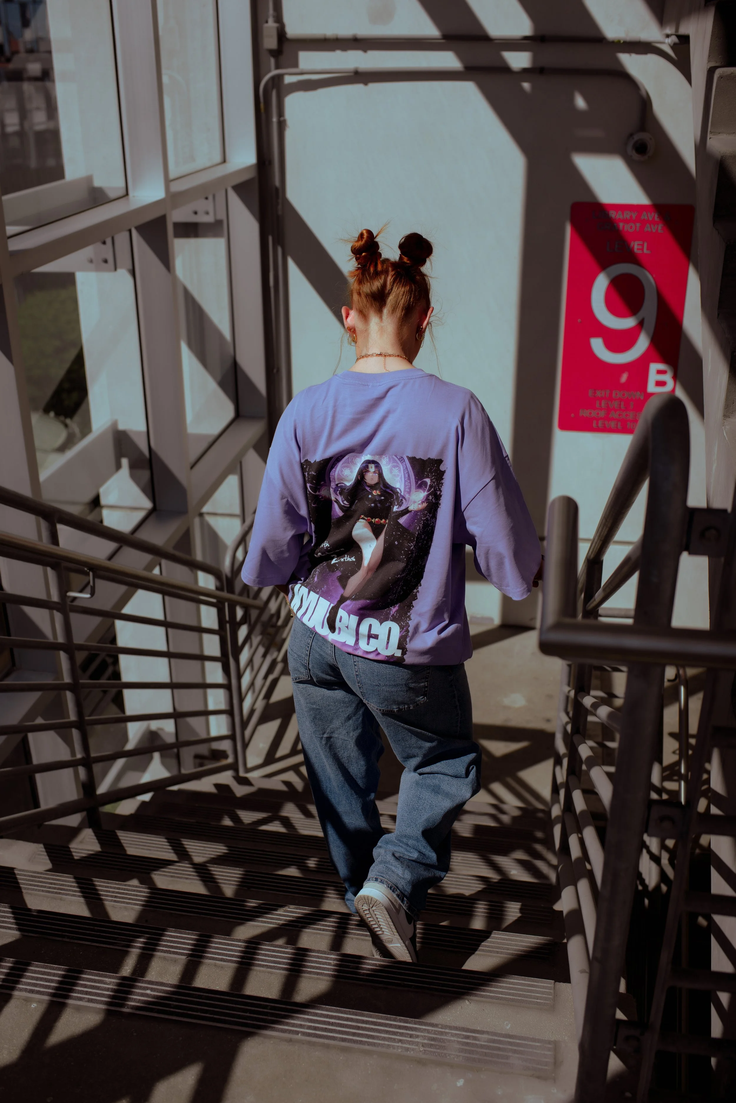 A woman with red hair styled in two buns, wearing a purple T-shirt with a graphic and the word 'MUBICO' on the back, and blue jeans, is walking down an outdoor staircase with metal railings, sunlight casting shadows on the stairs.