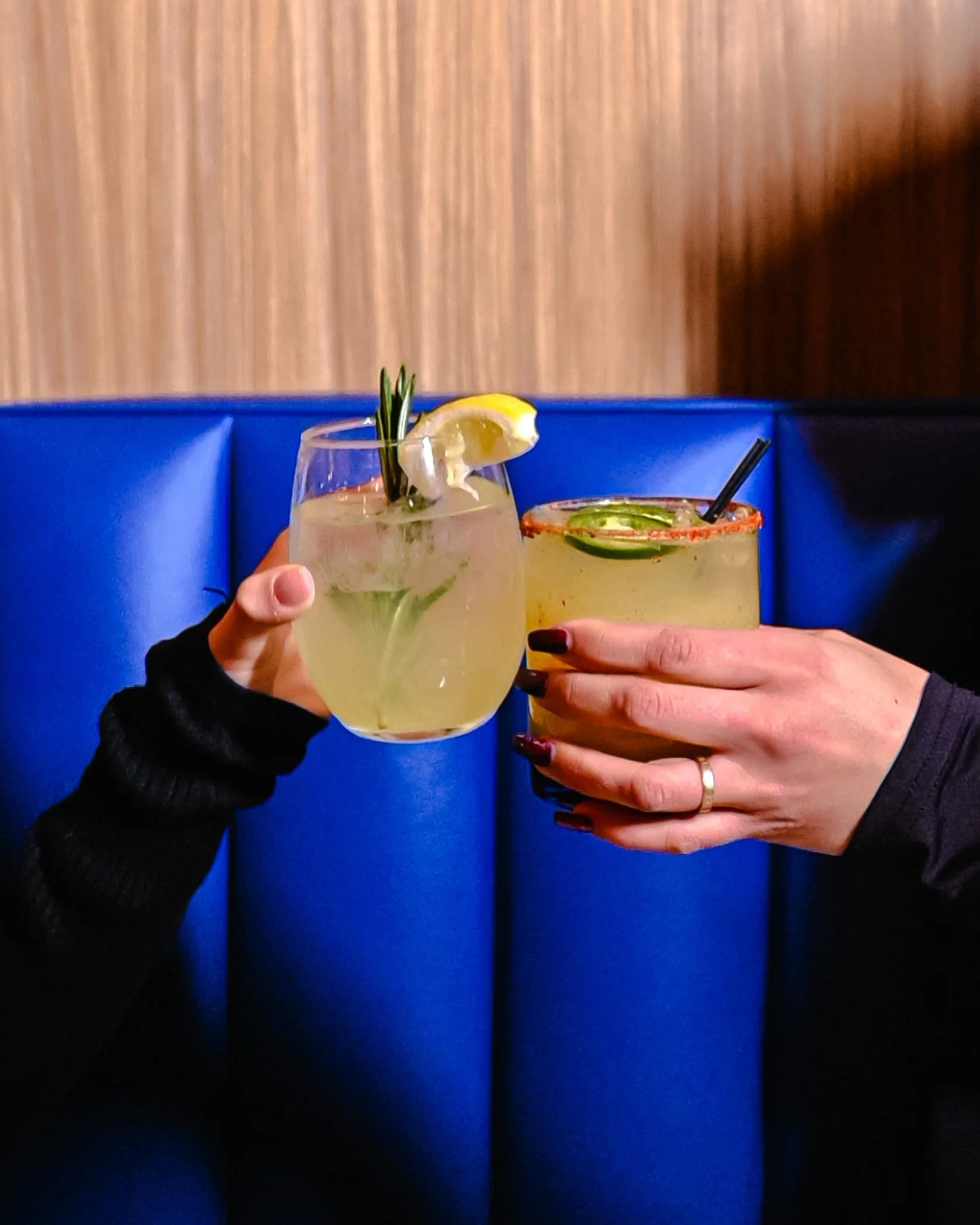 Two hands holding cocktails with garnishes, clinking in a toast