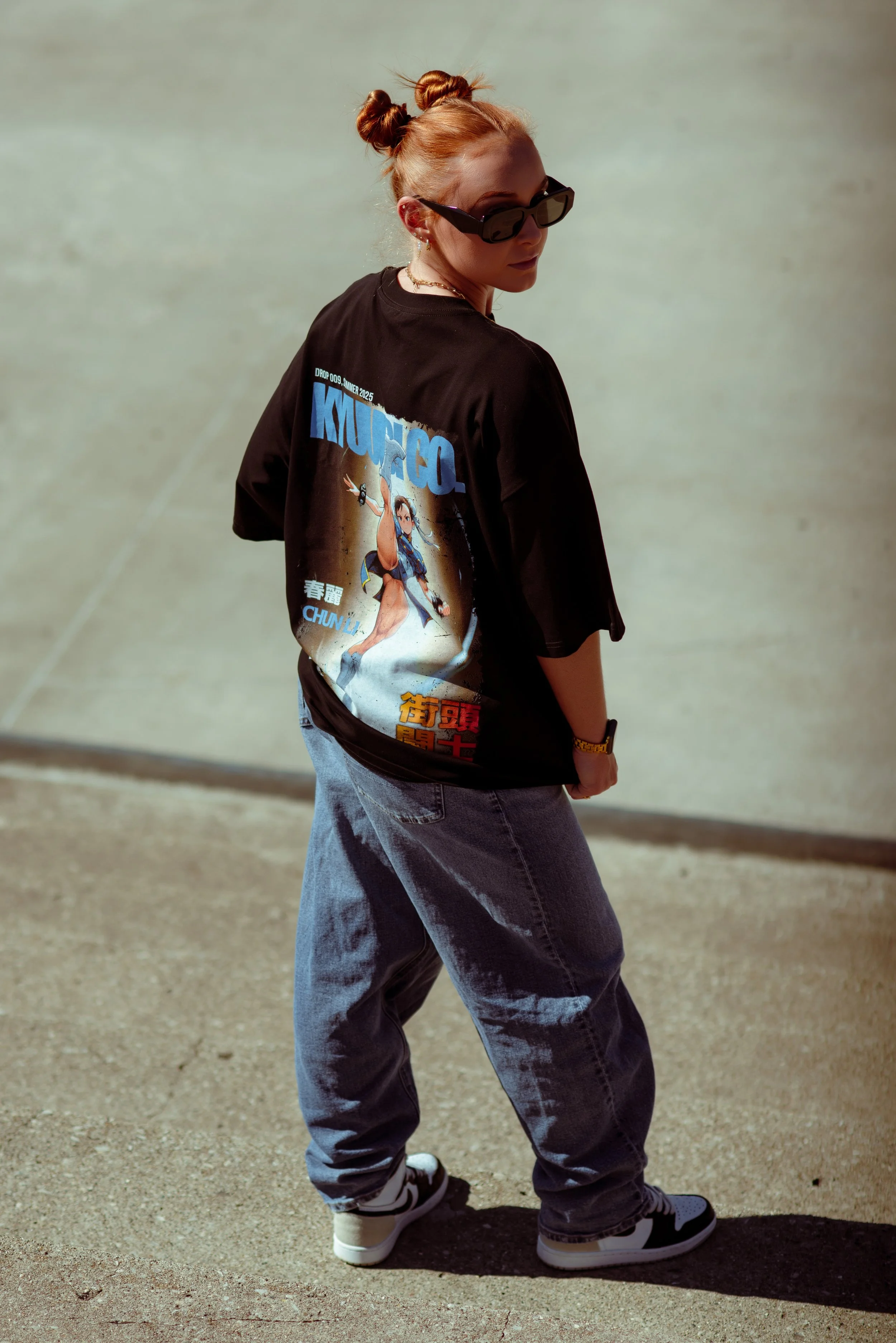 A young woman with red hair in two buns, wearing black sunglasses, a graphic T-shirt with anime-style artwork, baggy blue jeans, and sneakers, standing outdoors with a plain ground background.