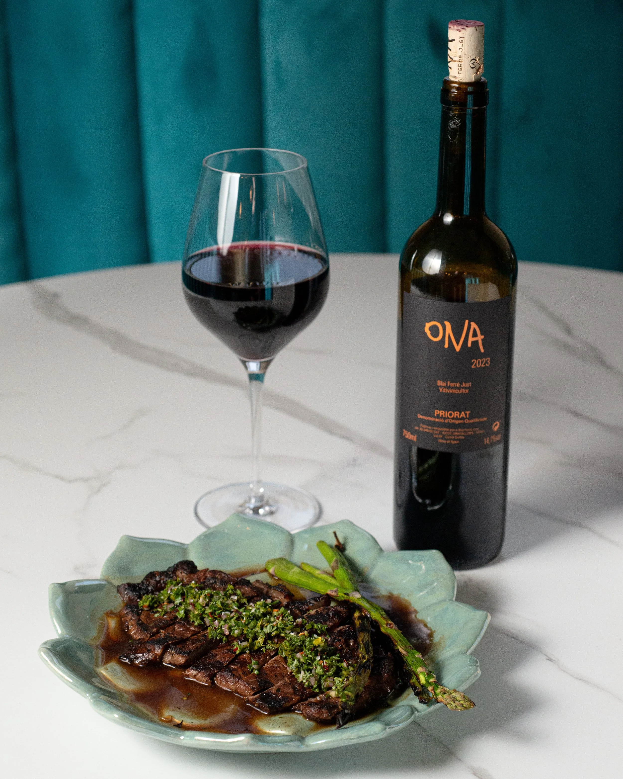 A glass of red wine, a bottle of ONA 2023 Priорат wine, and a plate of sliced beef with sauce and green garnish, along with a roasted green asparagus on a white marble table with a blue-green background.