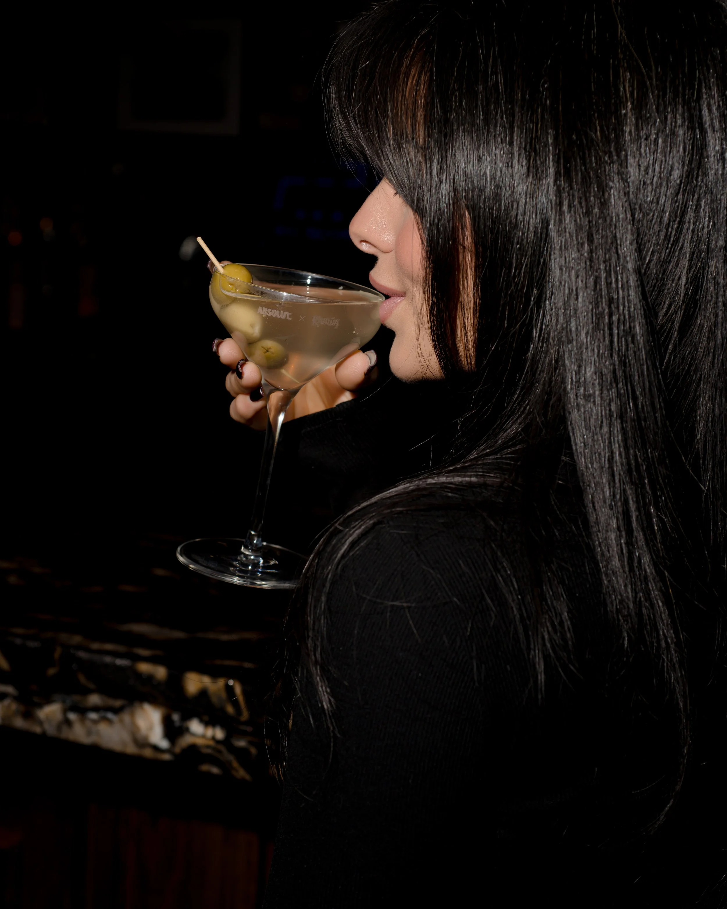 A woman with long black hair drinking a cocktail garnished with green olives.