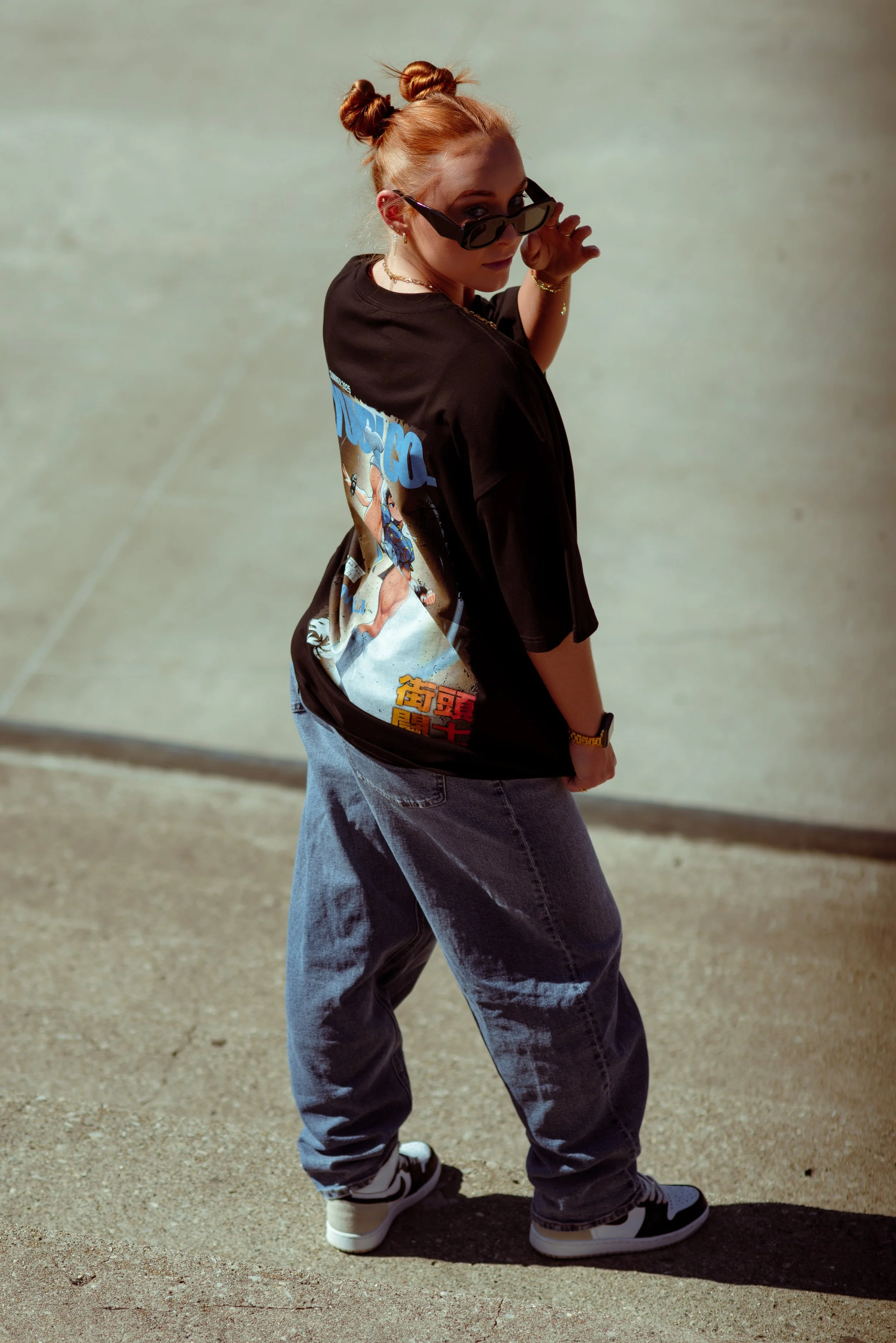 A young woman with red hair styled in two buns, wearing sunglasses, a black graphic t-shirt, loose jeans, and sneakers, posing outdoors on a sunny day.