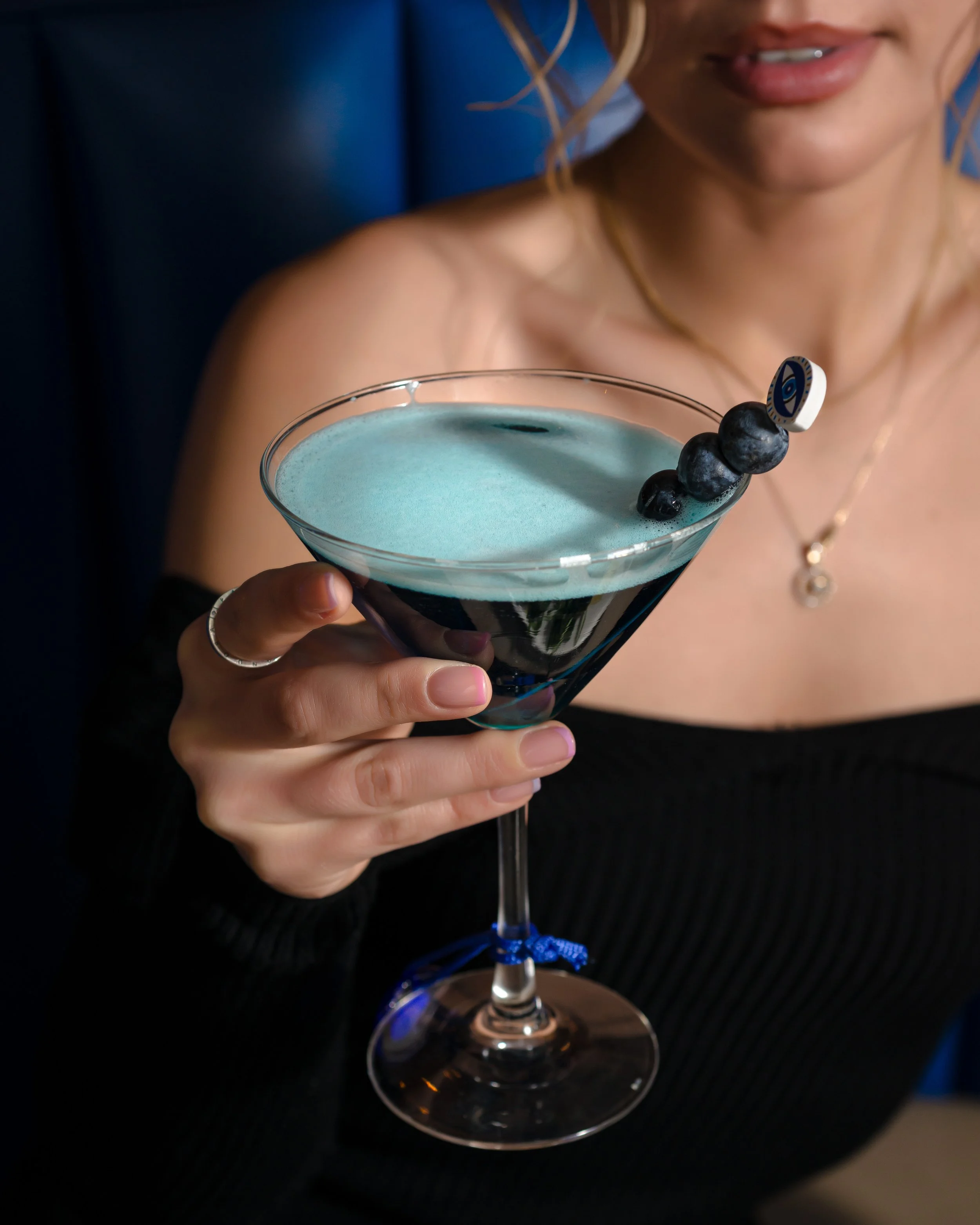 A woman holding a blue cocktail garnished with blueberries and a decorative eye-shaped pick, with her lips slightly parted.