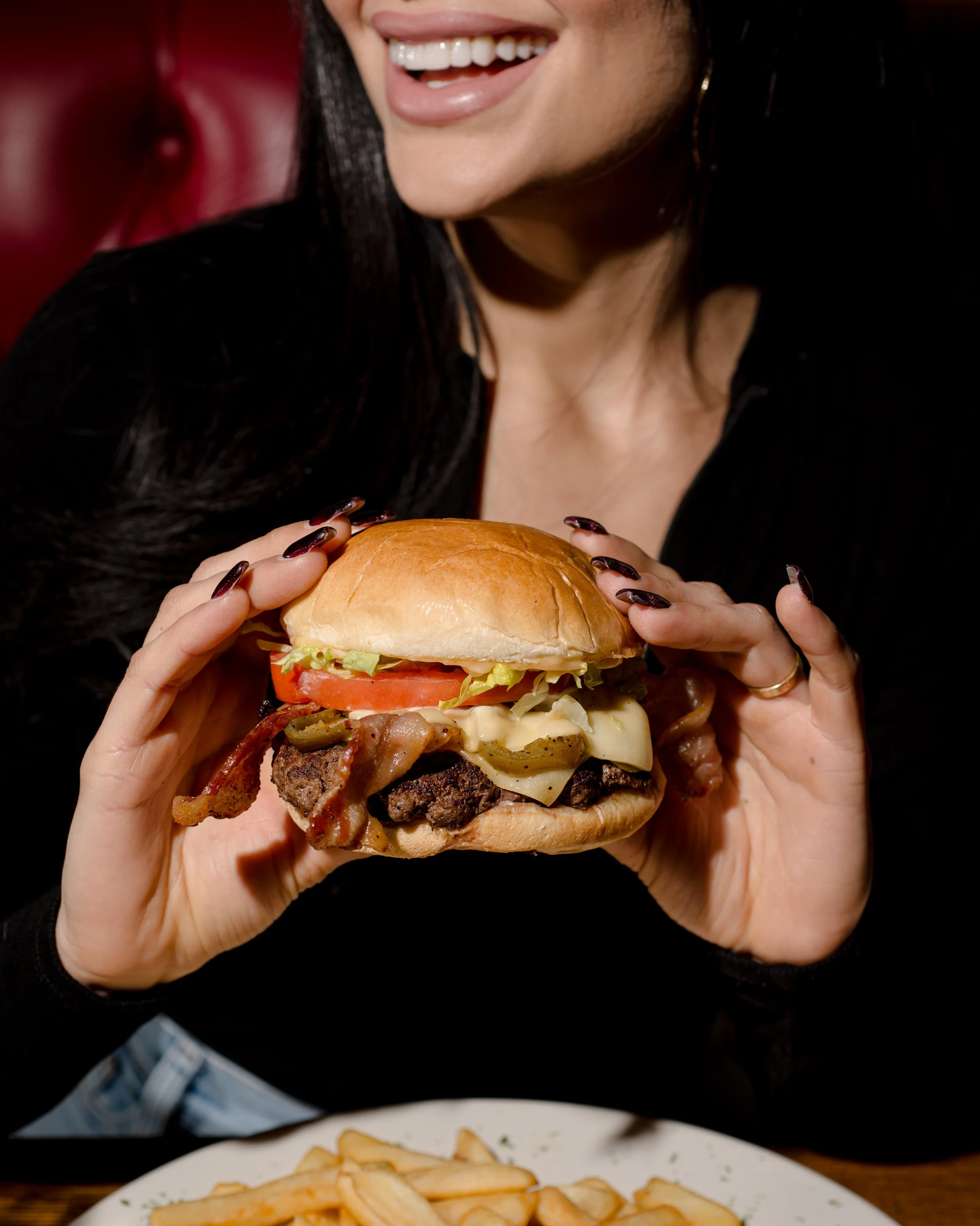 A woman holding a large burger with bacon, lettuce, tomato, cheese, and a beef patty, smiling with her teeth showing.