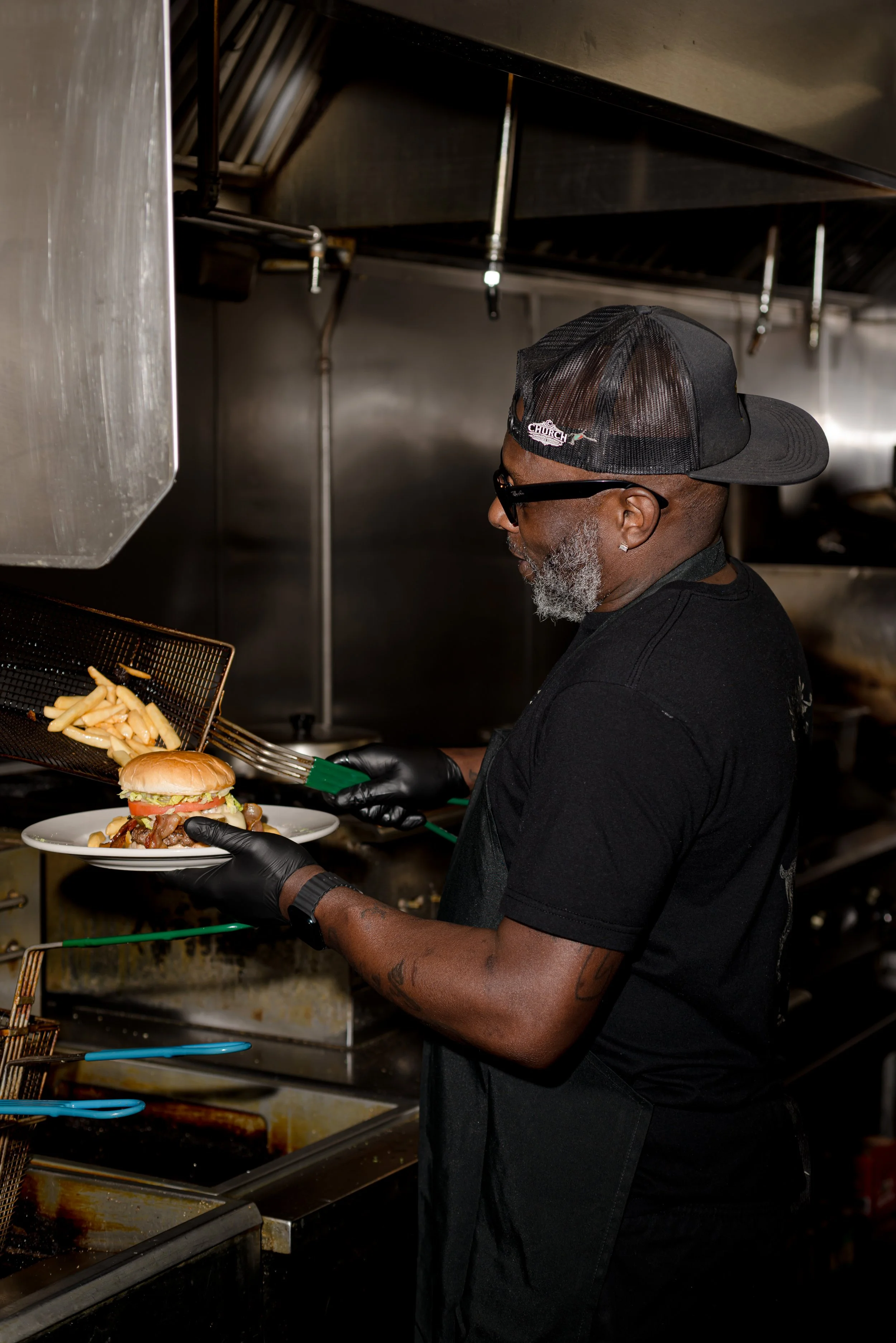 A man in a black cap, glasses, and black gloves preparing a burger with fries in a commercial kitchen.