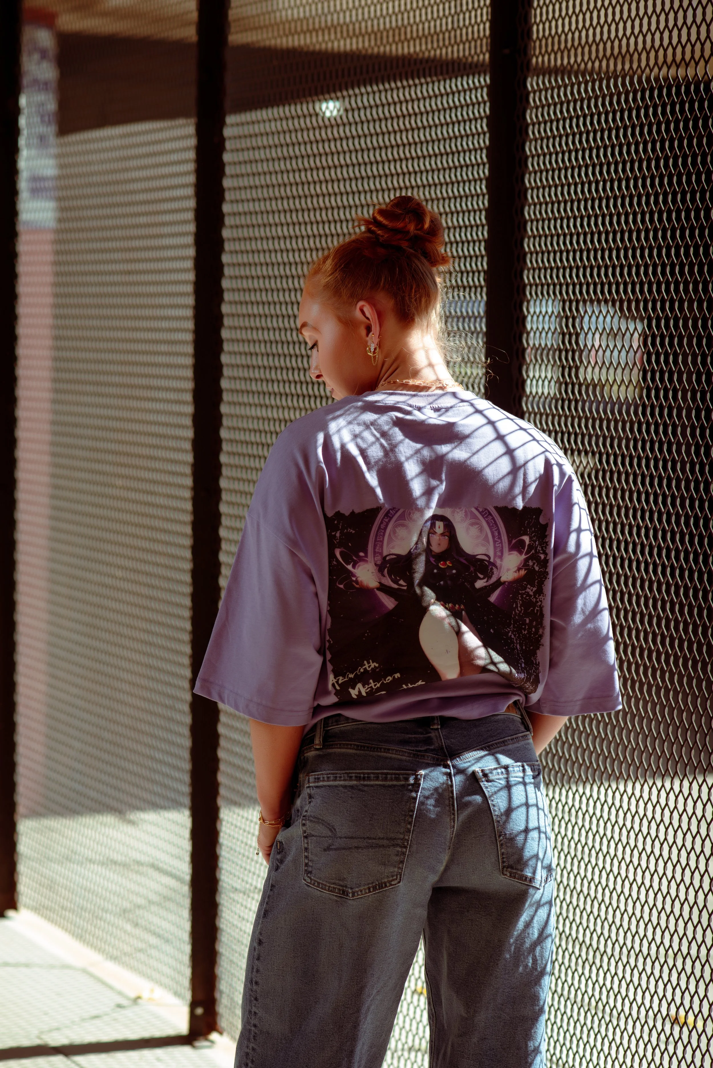 A woman with red hair tied in a bun stands with her back to the camera, lean against a metal mesh fence, wearing a lavender T-shirt with a graphic of a woman in black, and loose black jeans, casting shadows through the fence.