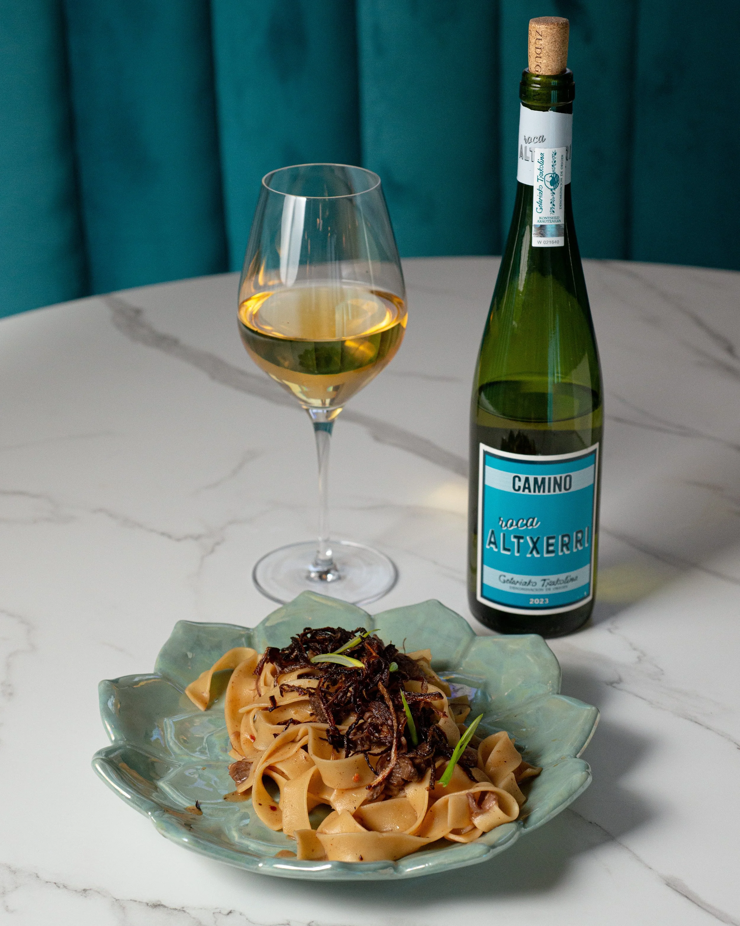 A tall glass of white wine, a bottle of white wine with a cork, and a plate of pasta with black truffle shavings on a white marble table.