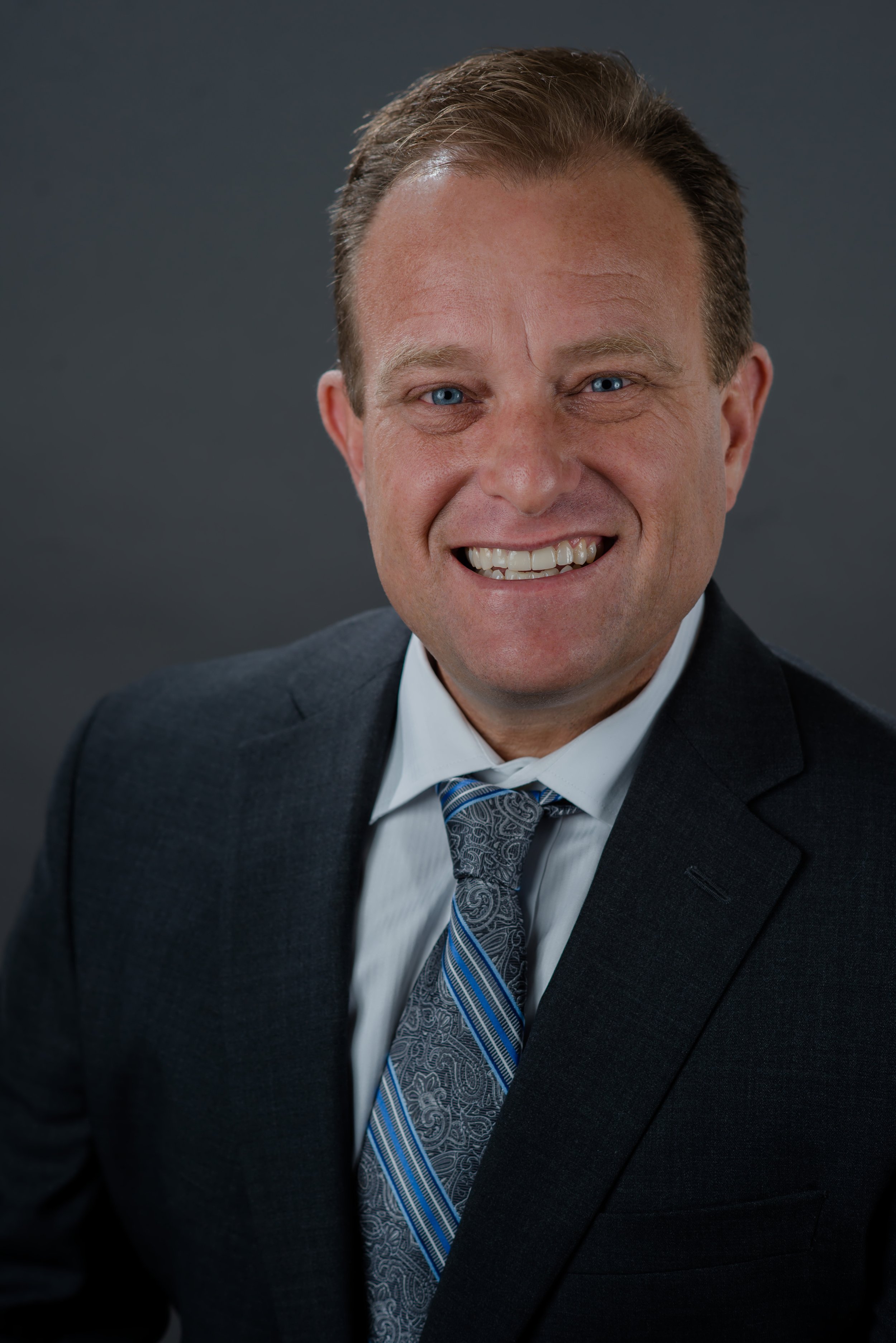 Close-up of a smiling man with blue eyes wearing a dark suit, white shirt, and patterned tie, set against a plain dark background.