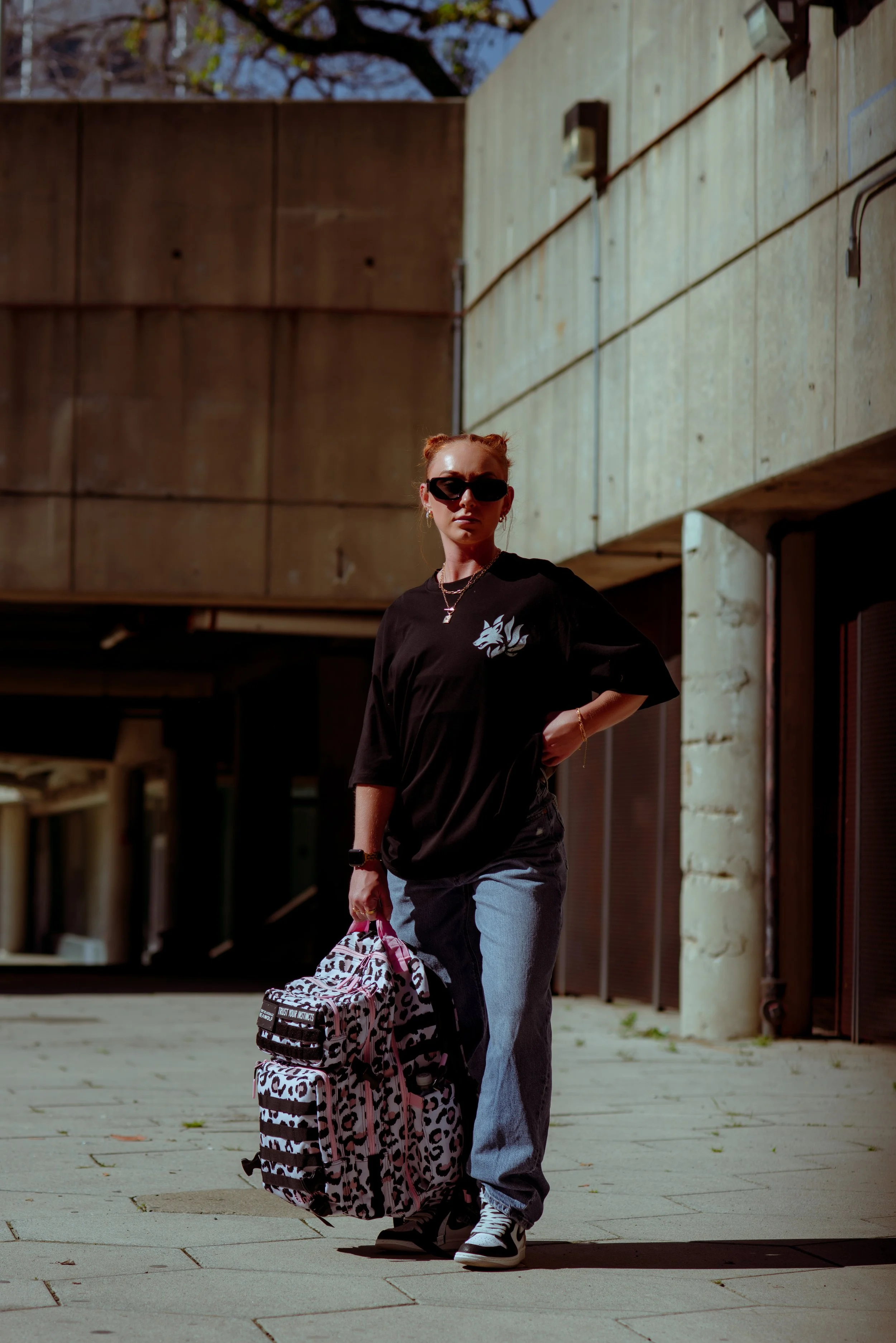 A young woman with red hair styled in buns wearing sunglasses, a black oversized t-shirt with a graphic, light blue jeans, and sneakers. She is carrying a leopard print backpack and walking outdoors in front of a modern concrete building.