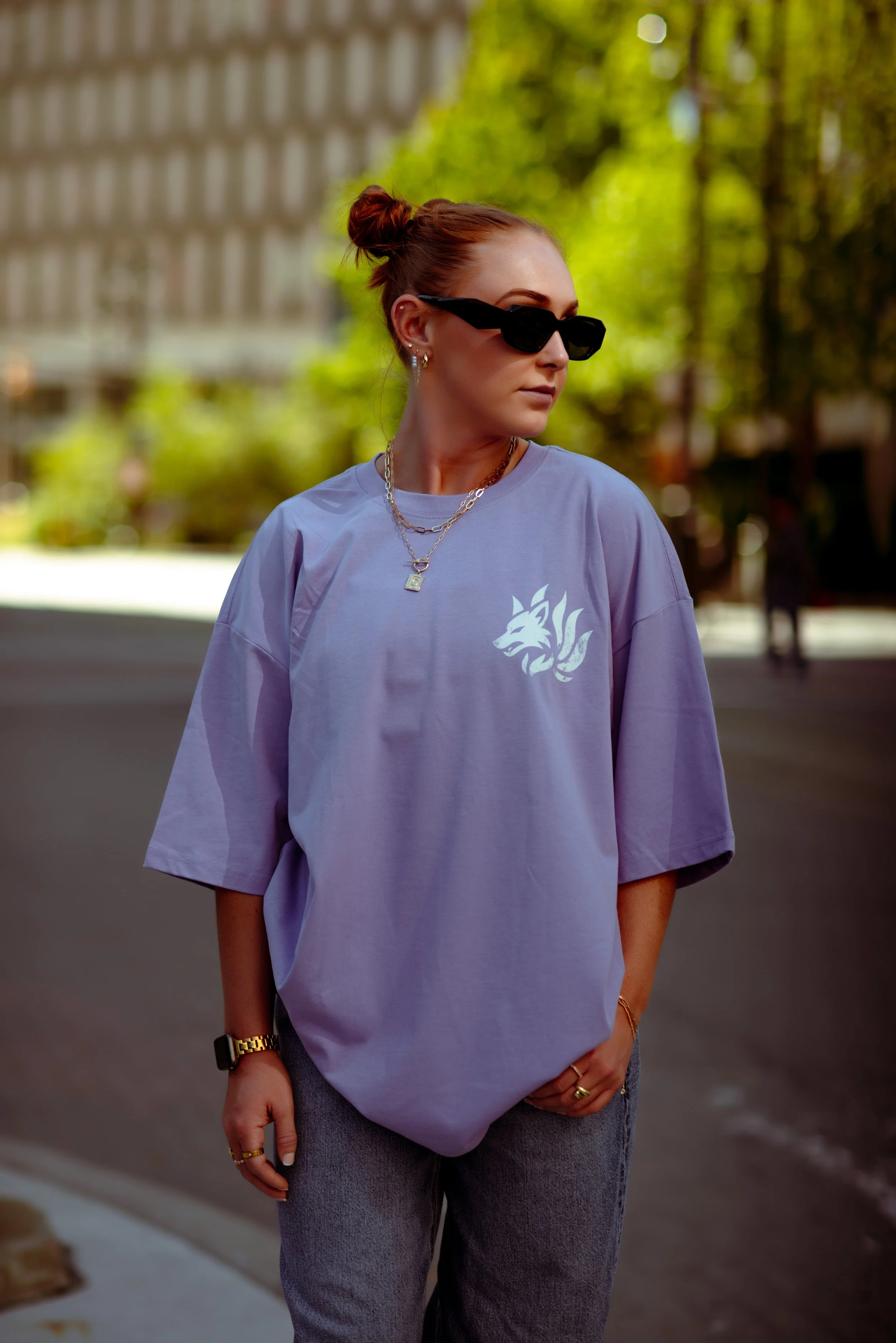A woman with red hair tied in a bun wearing black sunglasses, a loose lavender T-shirt with a wolf graphic, layered necklaces, and gray jeans, standing outdoors with trees and a building in the background.