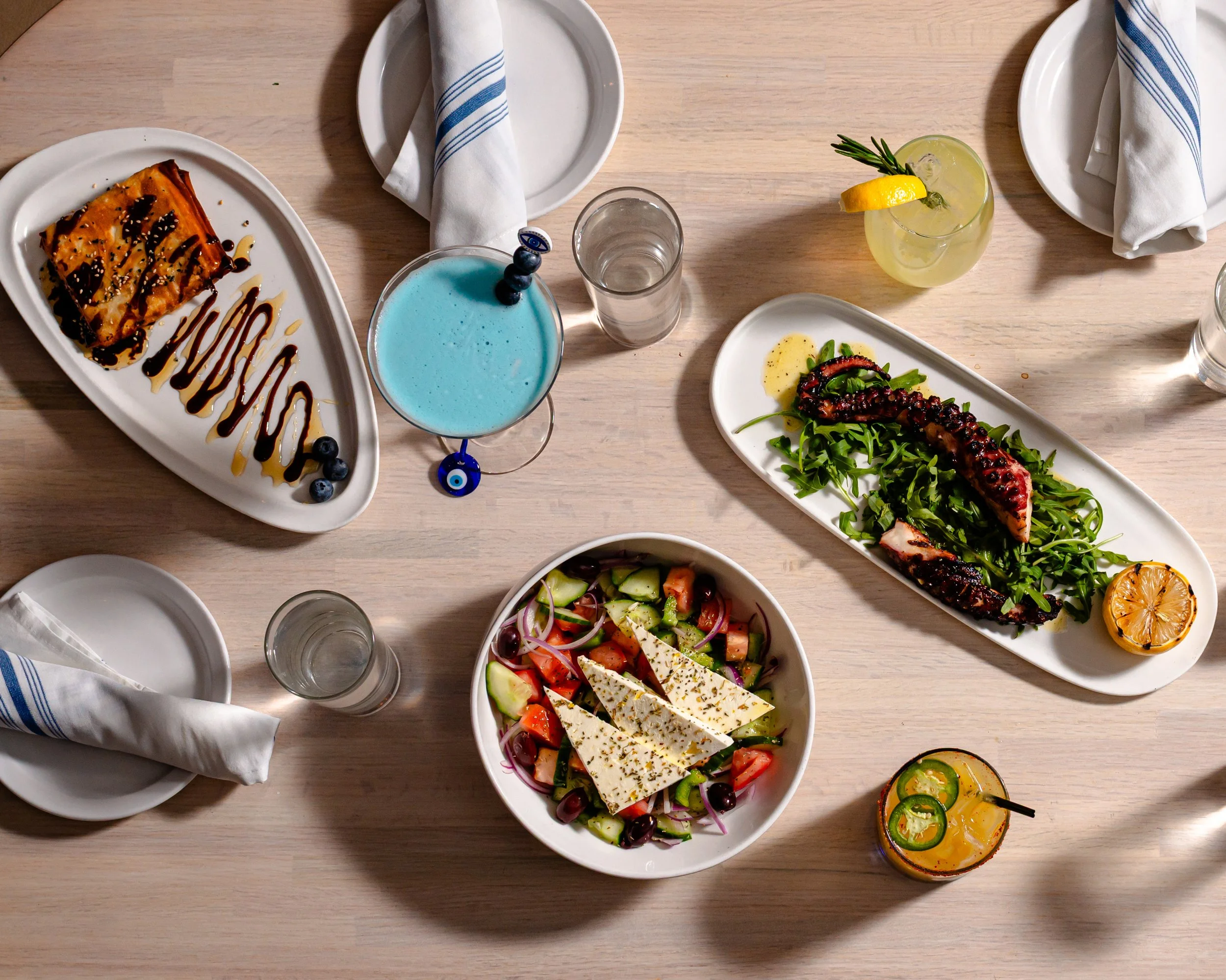 A top-down view of a table set with various dishes and drinks, including salad with feta cheese, grilled octopus with greens, a dessert with caramel sauce, and colorful cocktails.