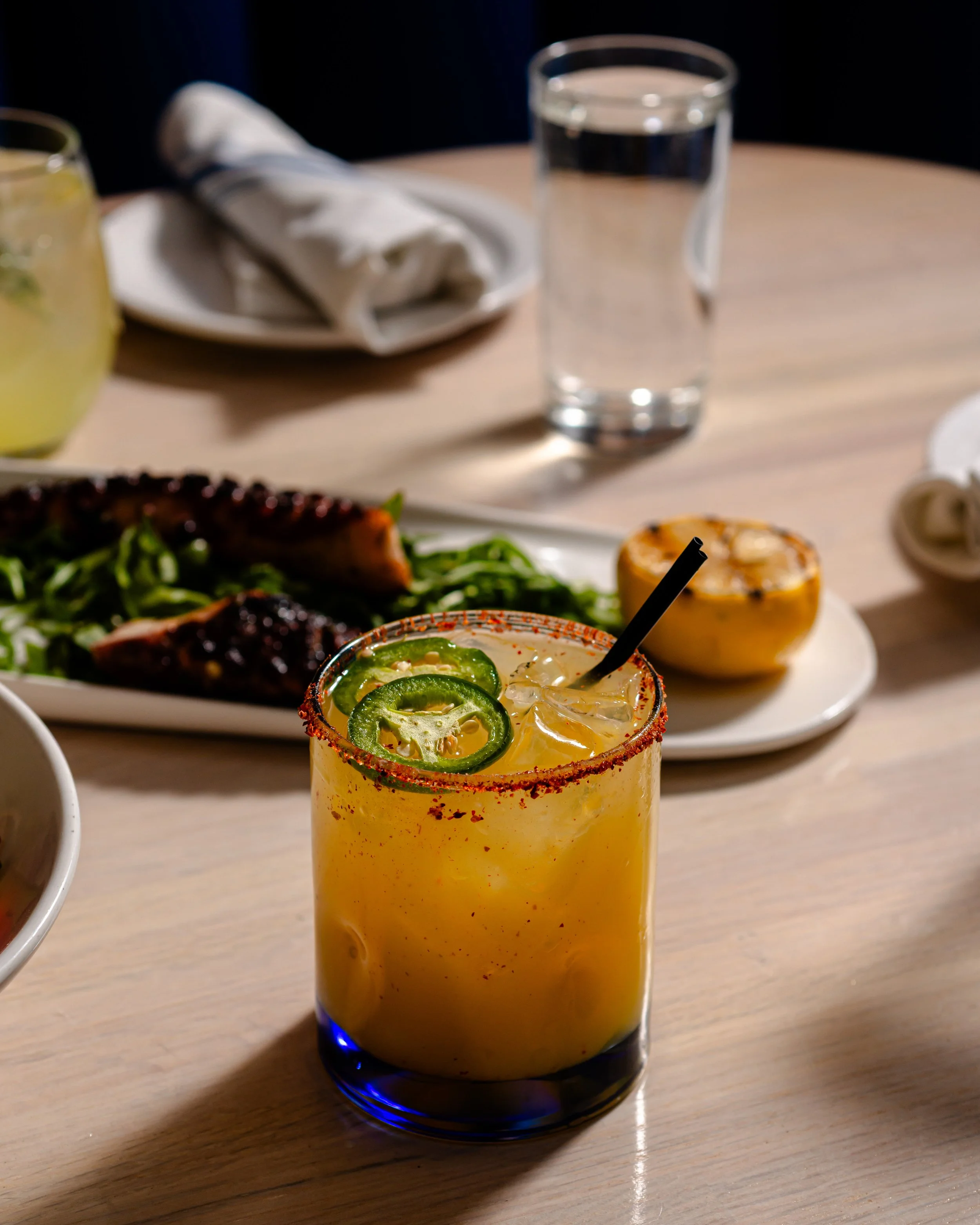 A yellow cocktail with jalapeño slices and a salted rim on a wooden table, with a plate of grilled meat, a half lemon, a glass of water, and a white napkin in the background.