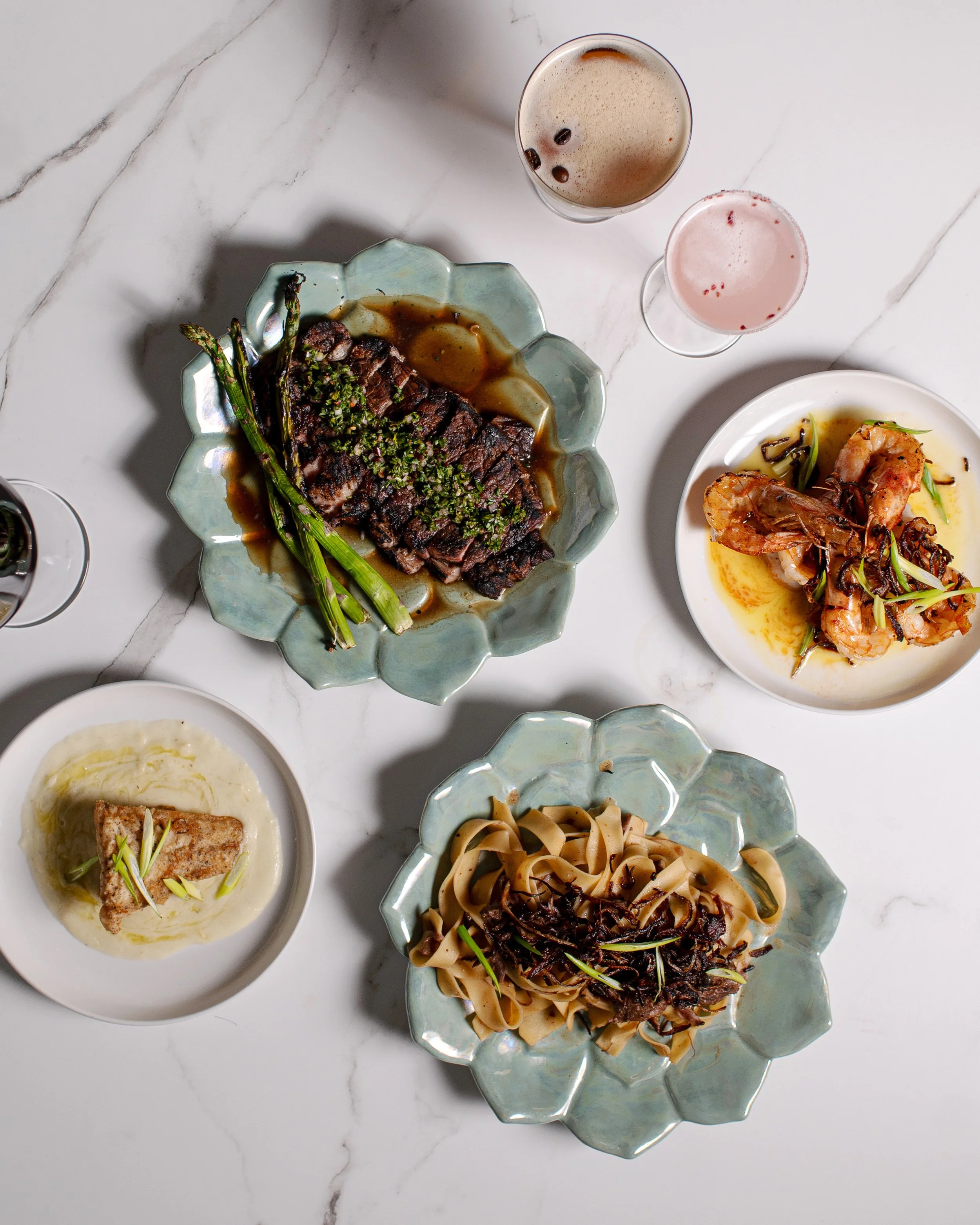 Table with various dishes including a plate of steak with green onions, a bowl of pasta with black sauce, a plate of shrimp, a small white dish with fish, and two glasses of beverages, on a white marble surface.