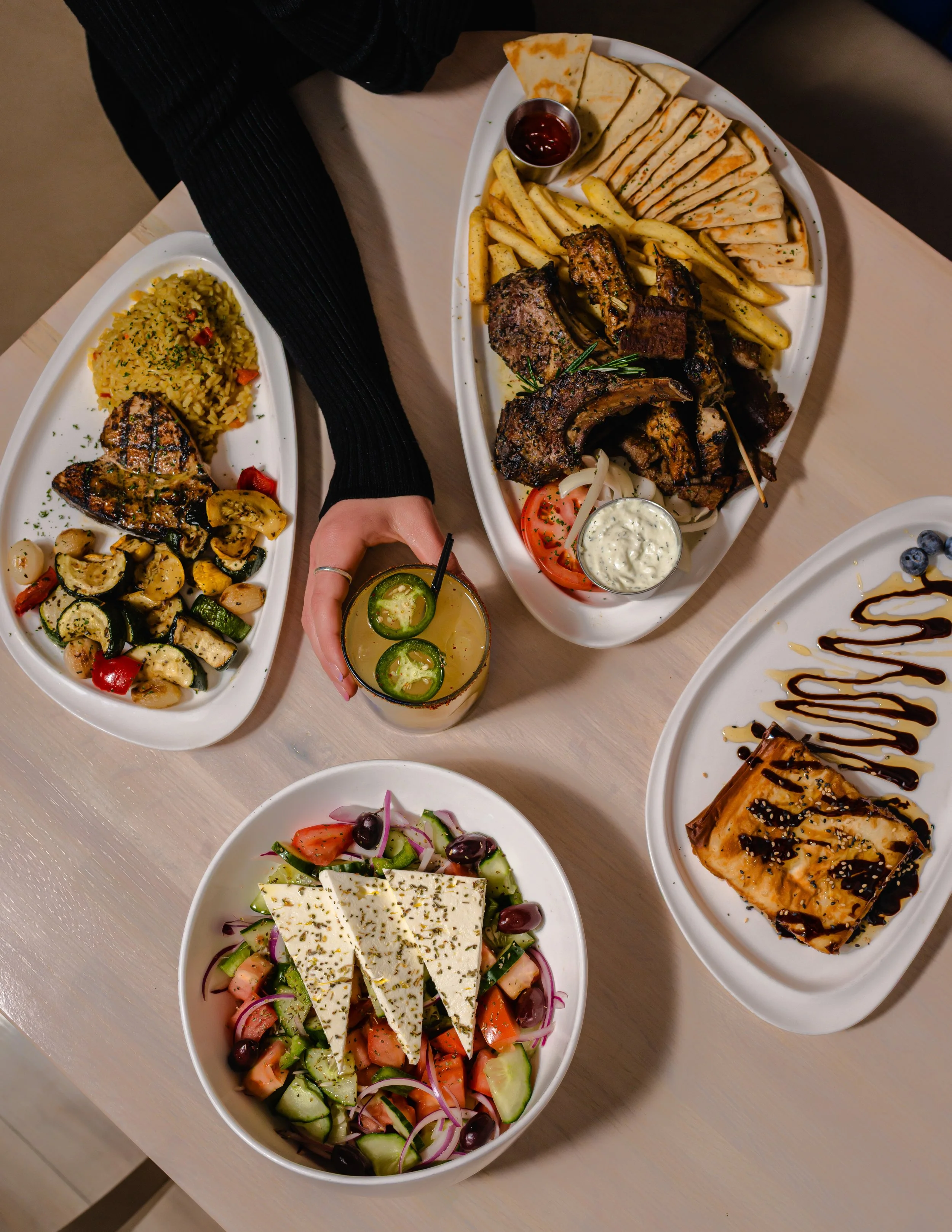 Top-down view of four plates of food, a person's hand holding a drink with jalapeno slices, on a light-colored wooden table.