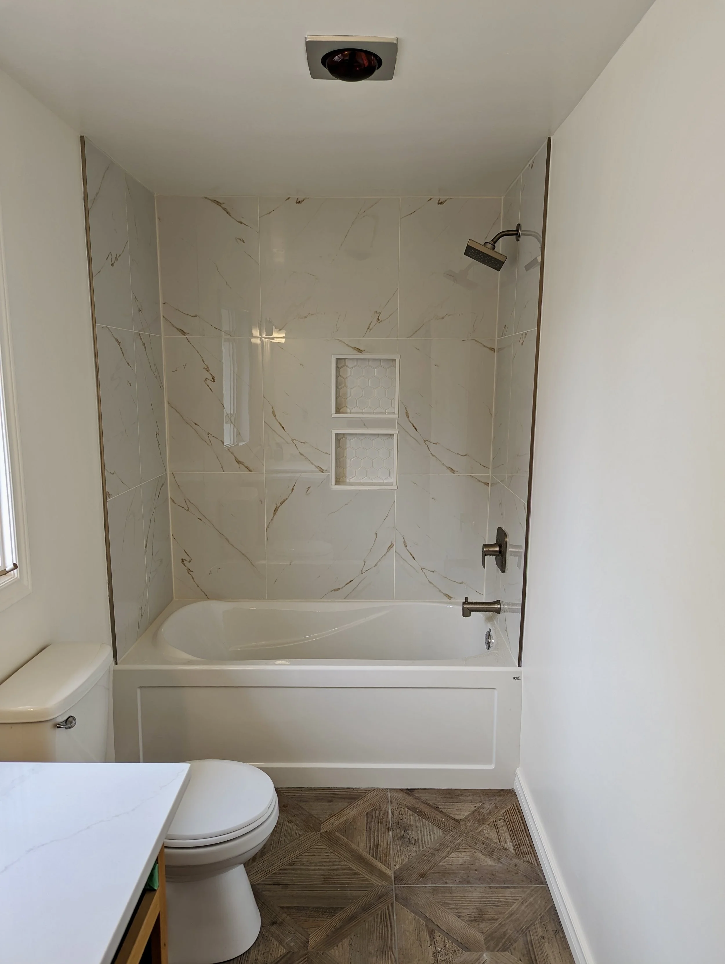 Bathroom with white bathtub, marble wall tiles, a showerhead, two built-in soap niches, a toilet, white walls, and wood-look tile flooring.