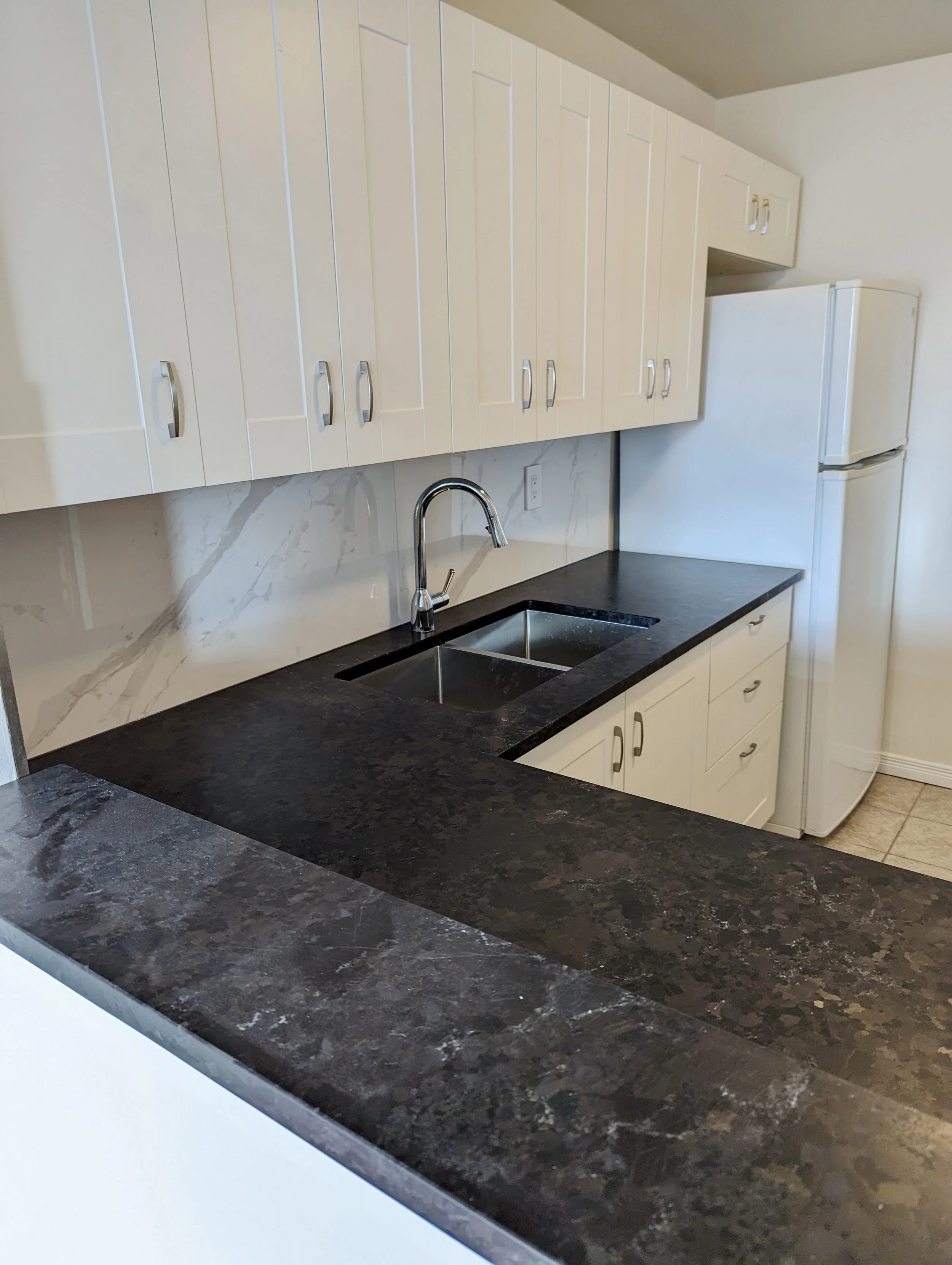 White kitchen with upper cabinets, black granite countertop, stainless steel sink with a chrome faucet, white refrigerator, and tiled floor.