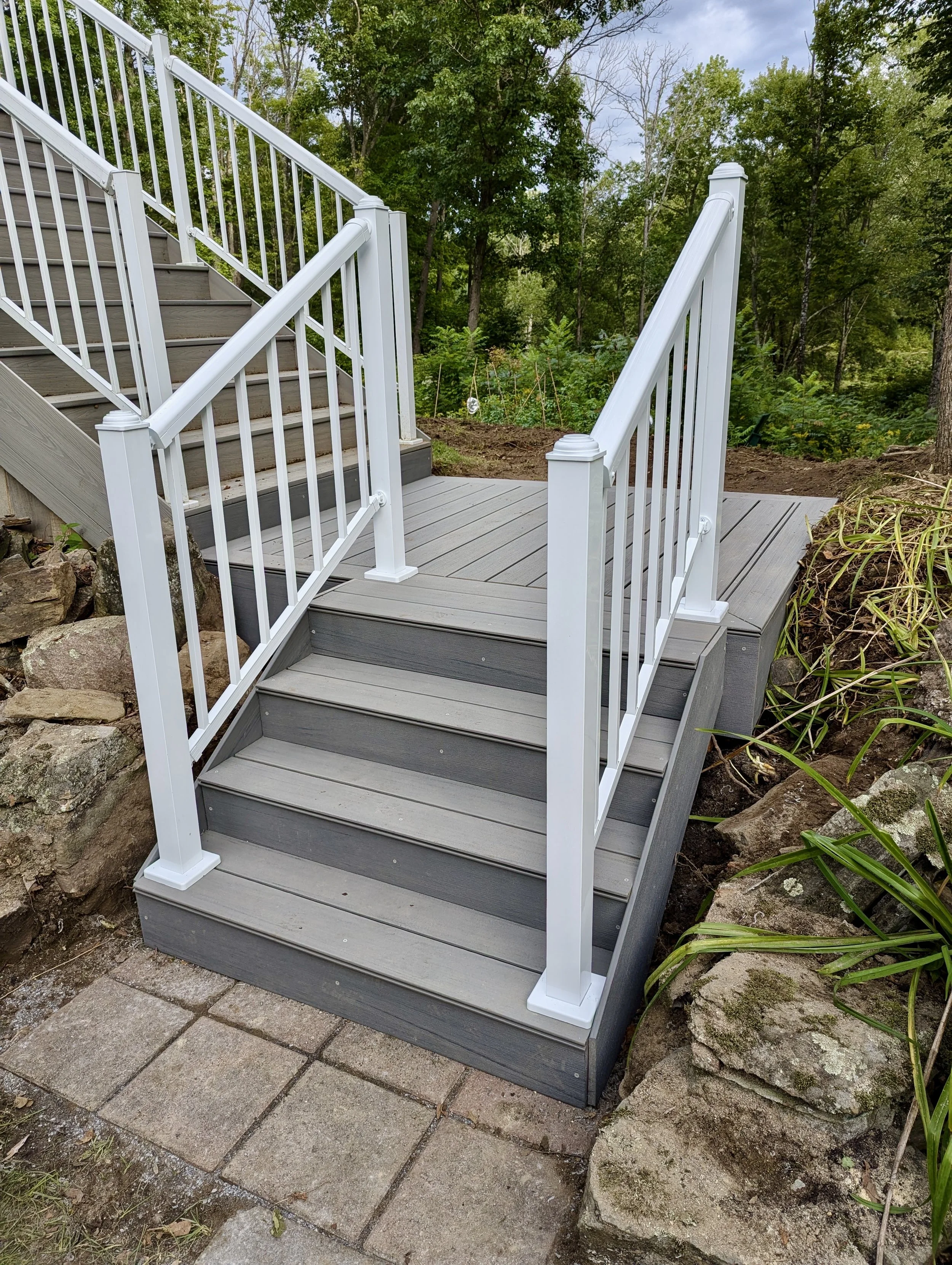 A newly built gray wooden deck with white metal railings and stairs leads down to a small paved area with stone pavers, surrounded by rocks and lush green trees.