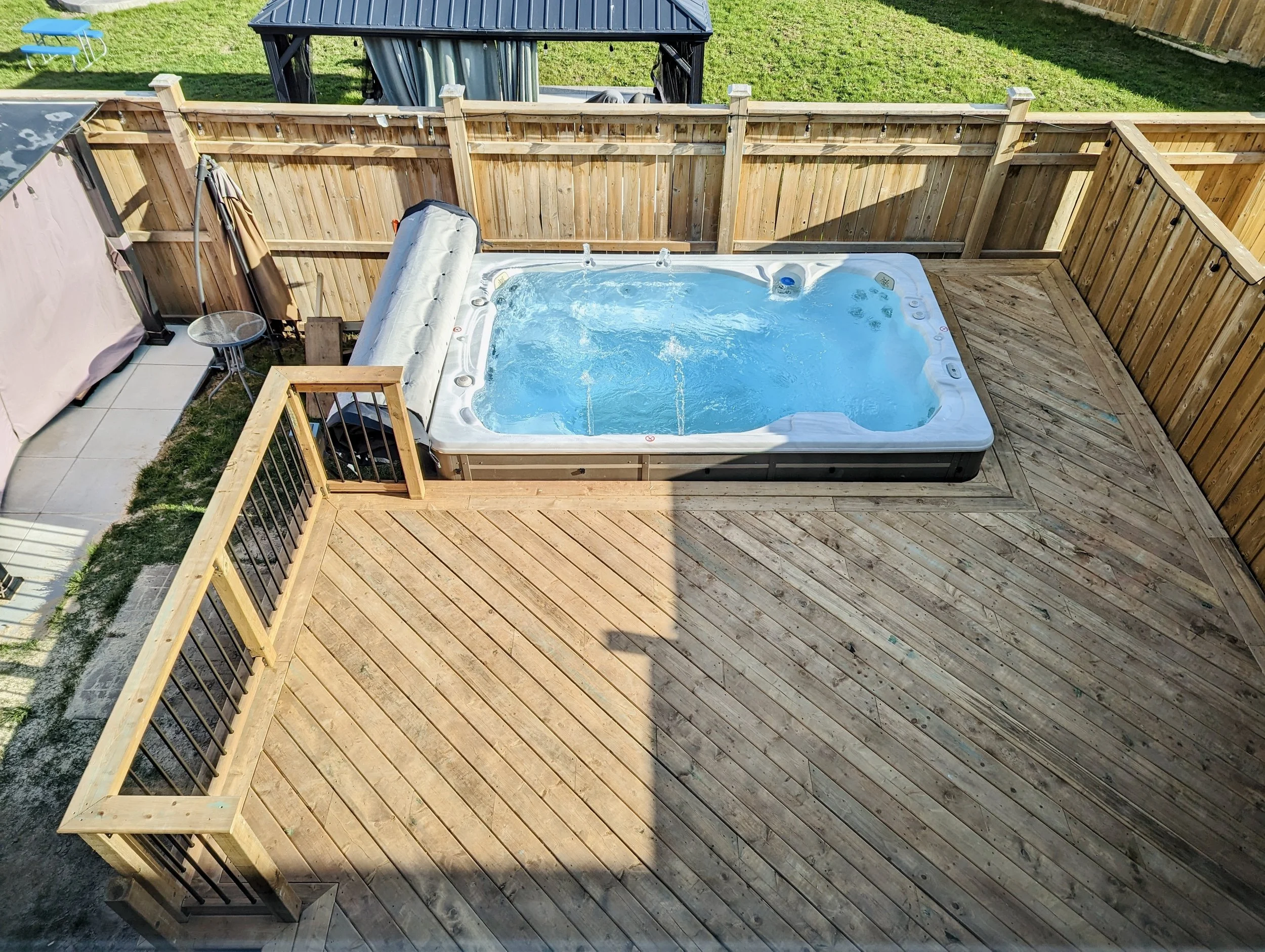 Backyard wooden deck with a hot tub filled with water, enclosed by a wooden privacy fence. A shaded area with a small round table and a covered grill. The lawn with grass and a shed or storage structure visible in the background.