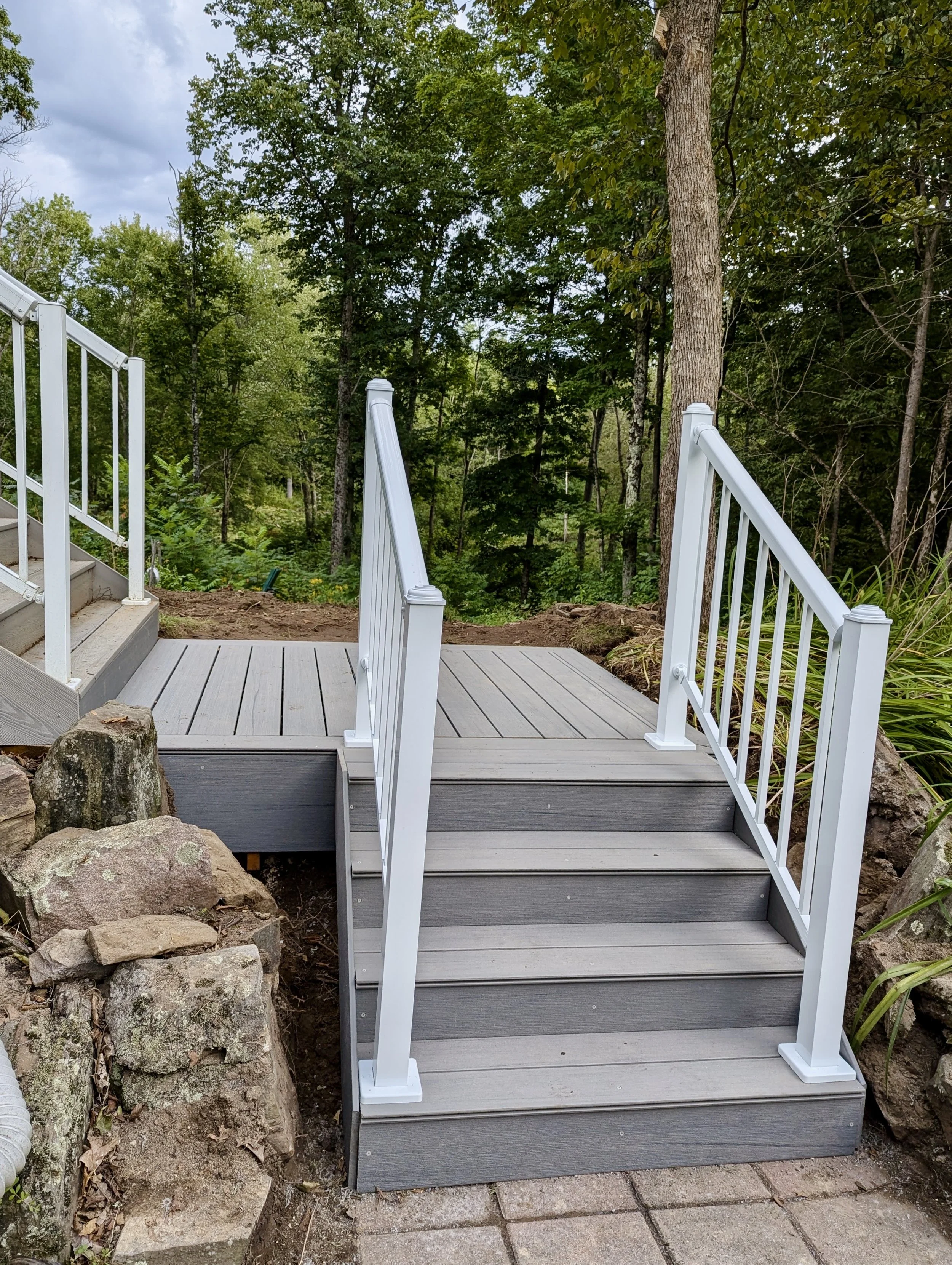 Small outdoor wooden deck with white railing leads to a rustic area with rocks, surrounded by green trees and forest.