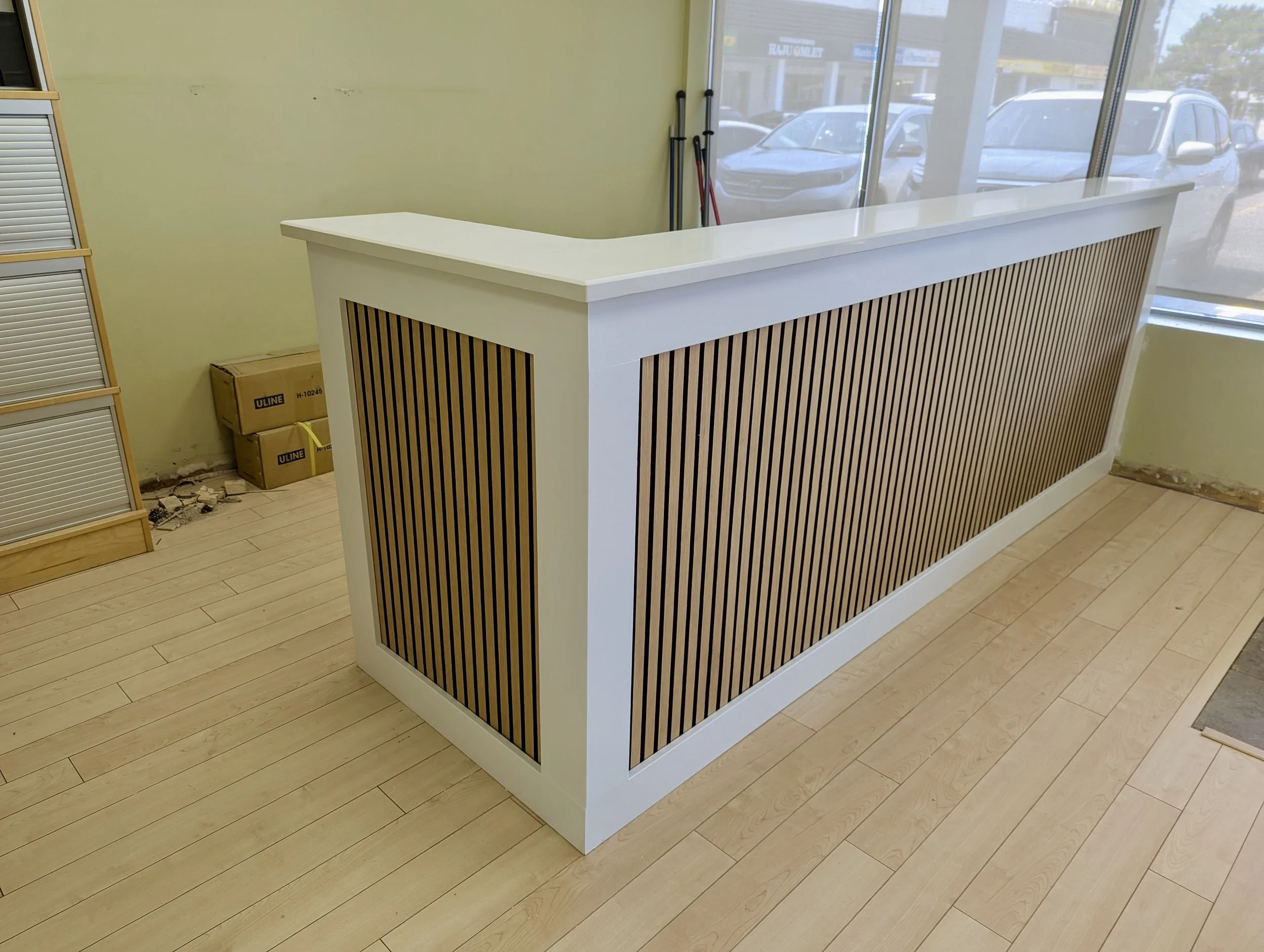 White reception counter with wooden slat panels in a retail or office space, with a large glass window showing parked cars outside.