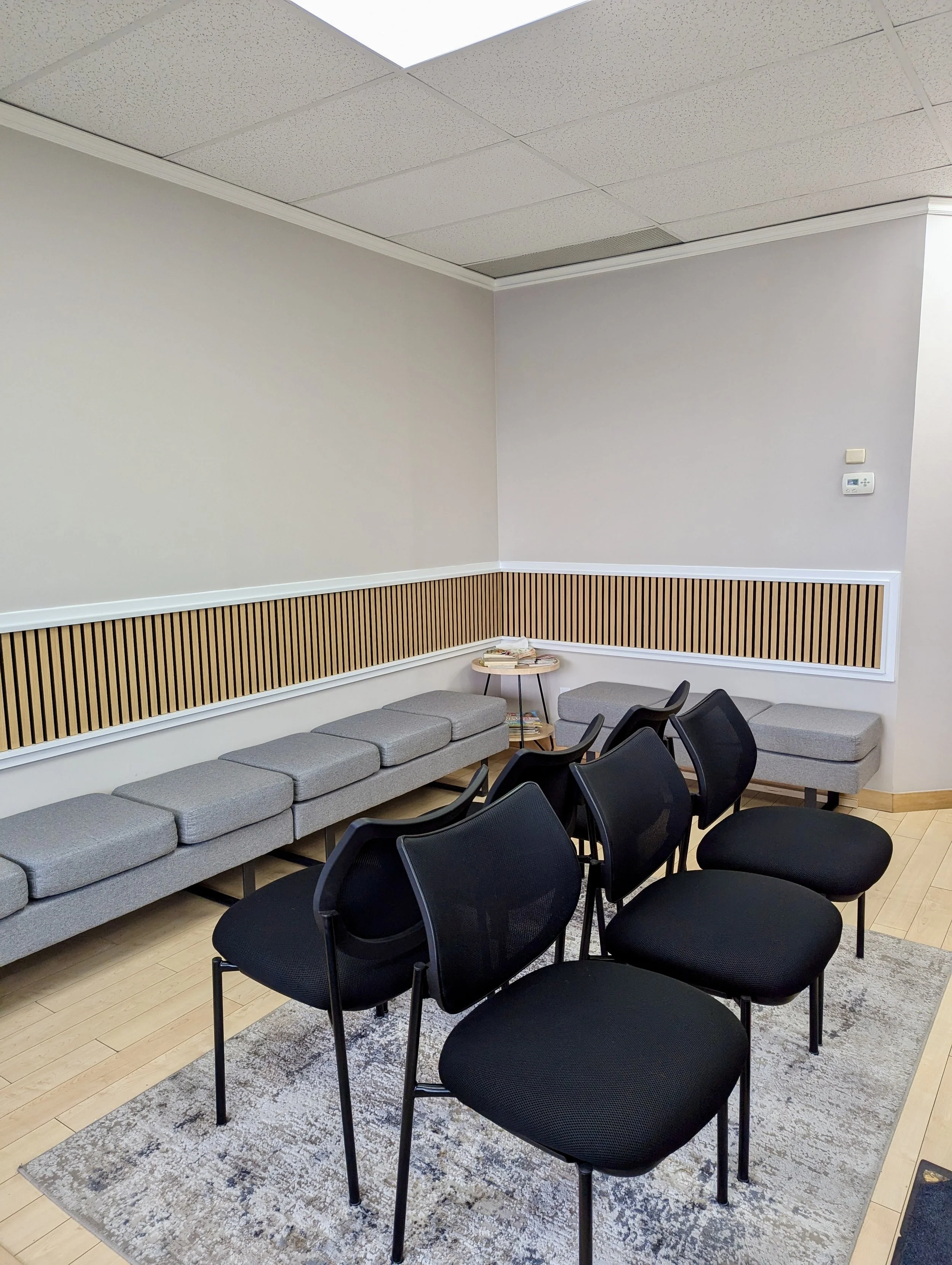 A small waiting room with six black chairs, a gray upholstered bench, a beige area rug, a corner table with magazines, and a wood-paneled wall.