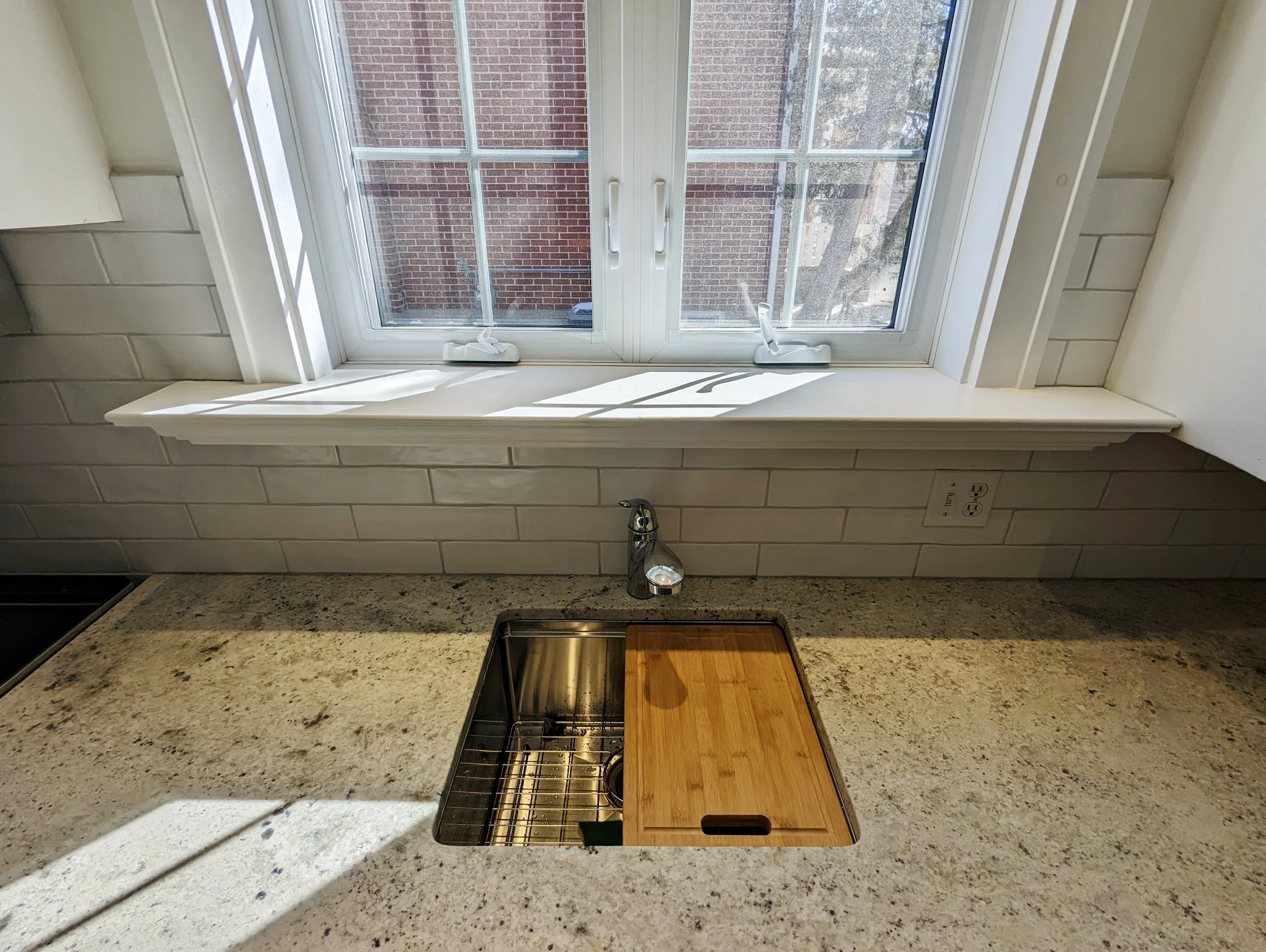 Kitchen countertop with a stainless steel sink, a wooden cutting board covering part of the sink, and a window above the counter showing brick buildings outside.