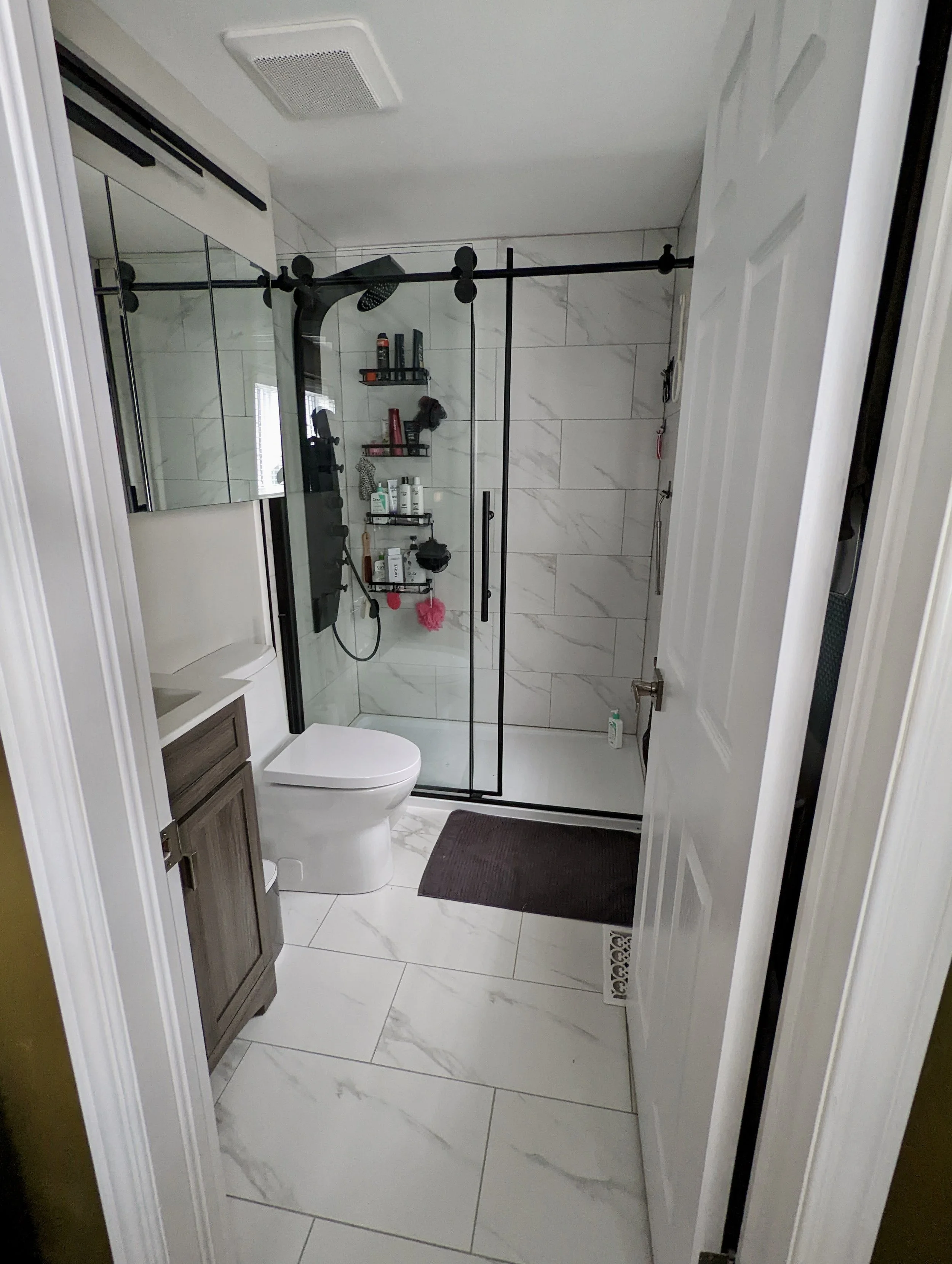 Modern bathroom with a glass-enclosed shower, white marble tiles, a toilet, a wooden vanity with a sink, and black metal shelving inside the shower holding toiletries.