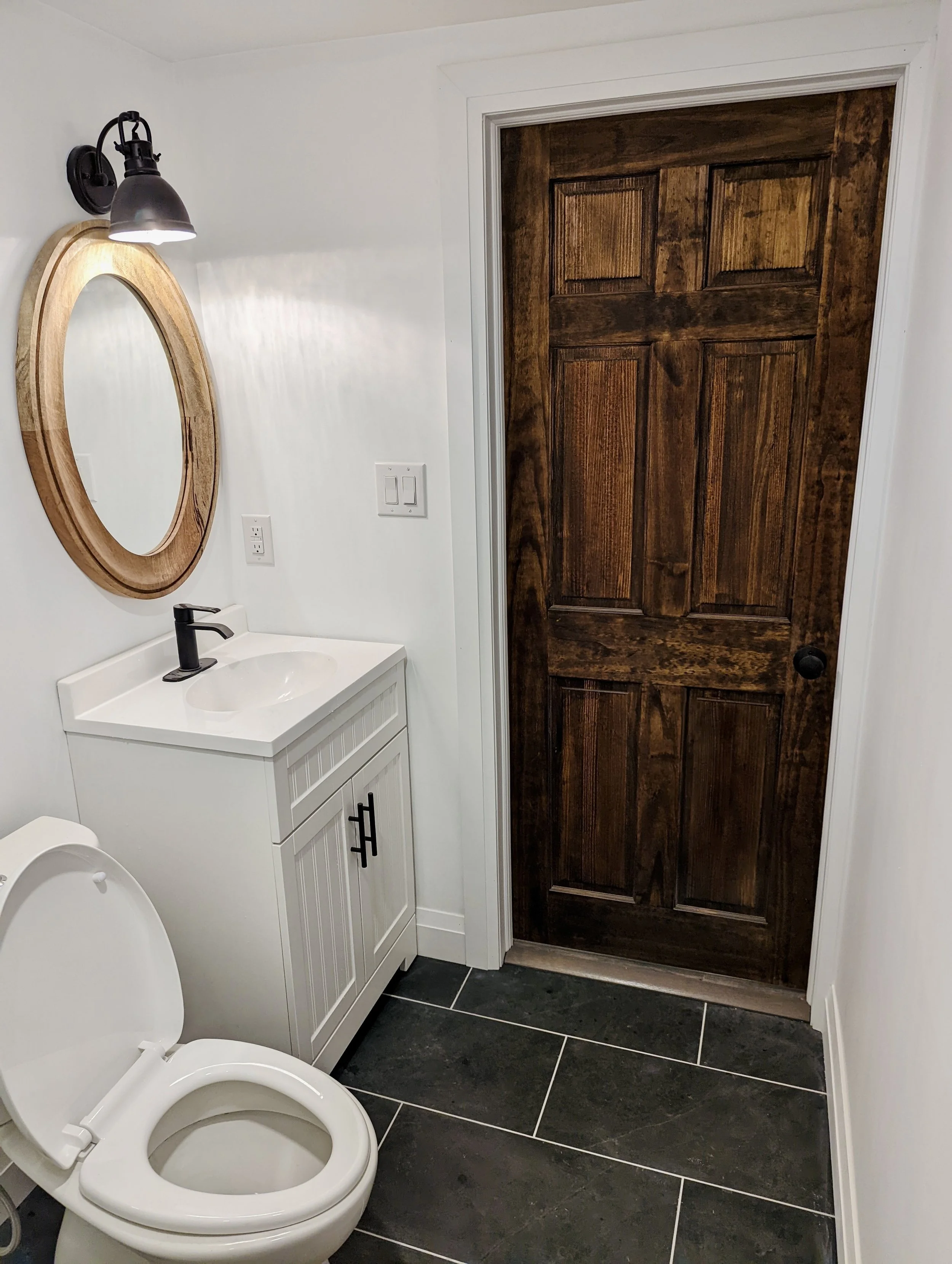 Bathroom with white walls, a black light fixture, a round wooden mirror, a white vanity with a black faucet, a white toilet, and a dark wooden door.