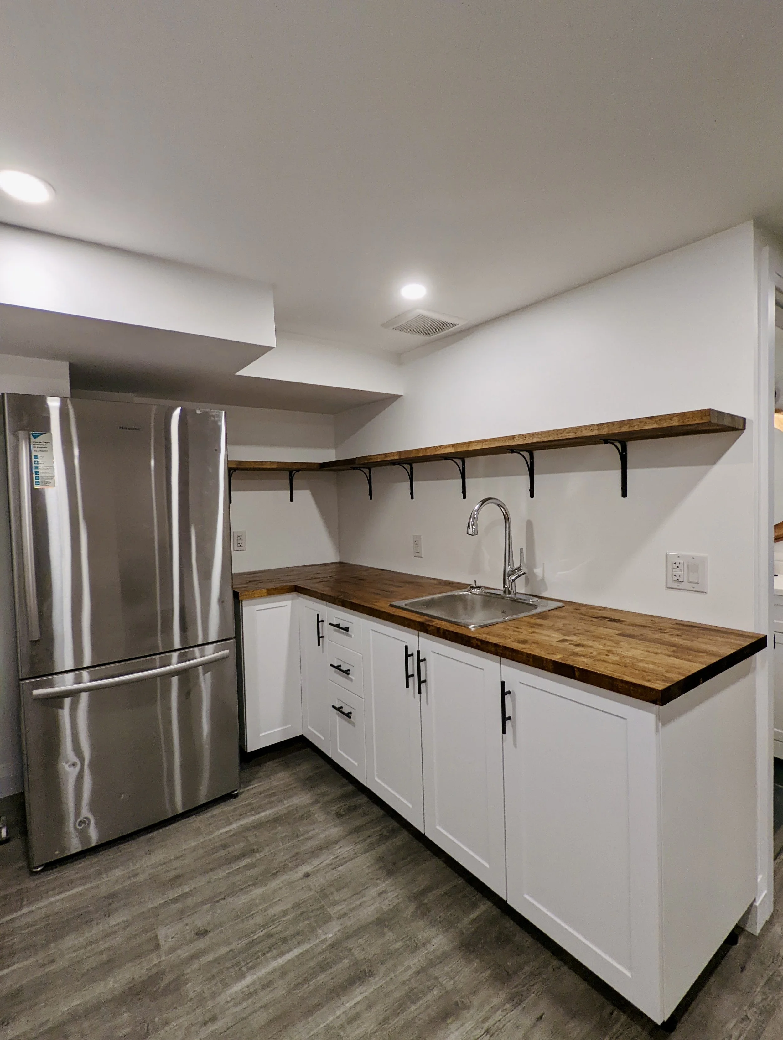 Kitchen with white cabinets, wooden countertop, stainless steel refrigerator, open wooden shelf, white walls, dark wood flooring, and a sink.