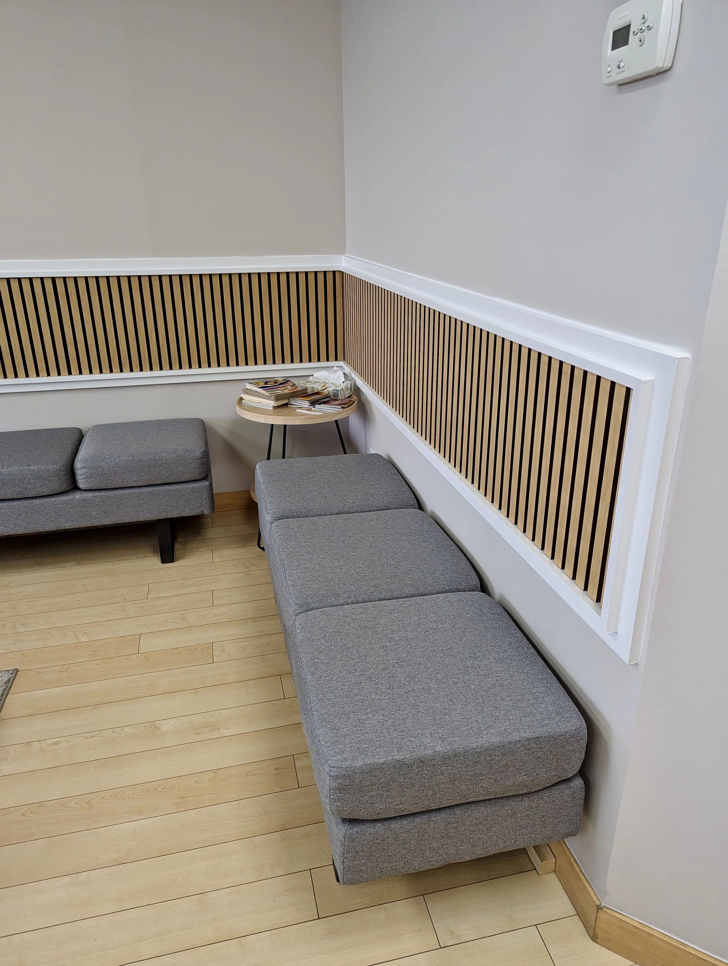 A gray upholstered seating area with a small round wooden table, magazines, and snacks in a minimalist room with wooden flooring and a wooden slat wall accent.
