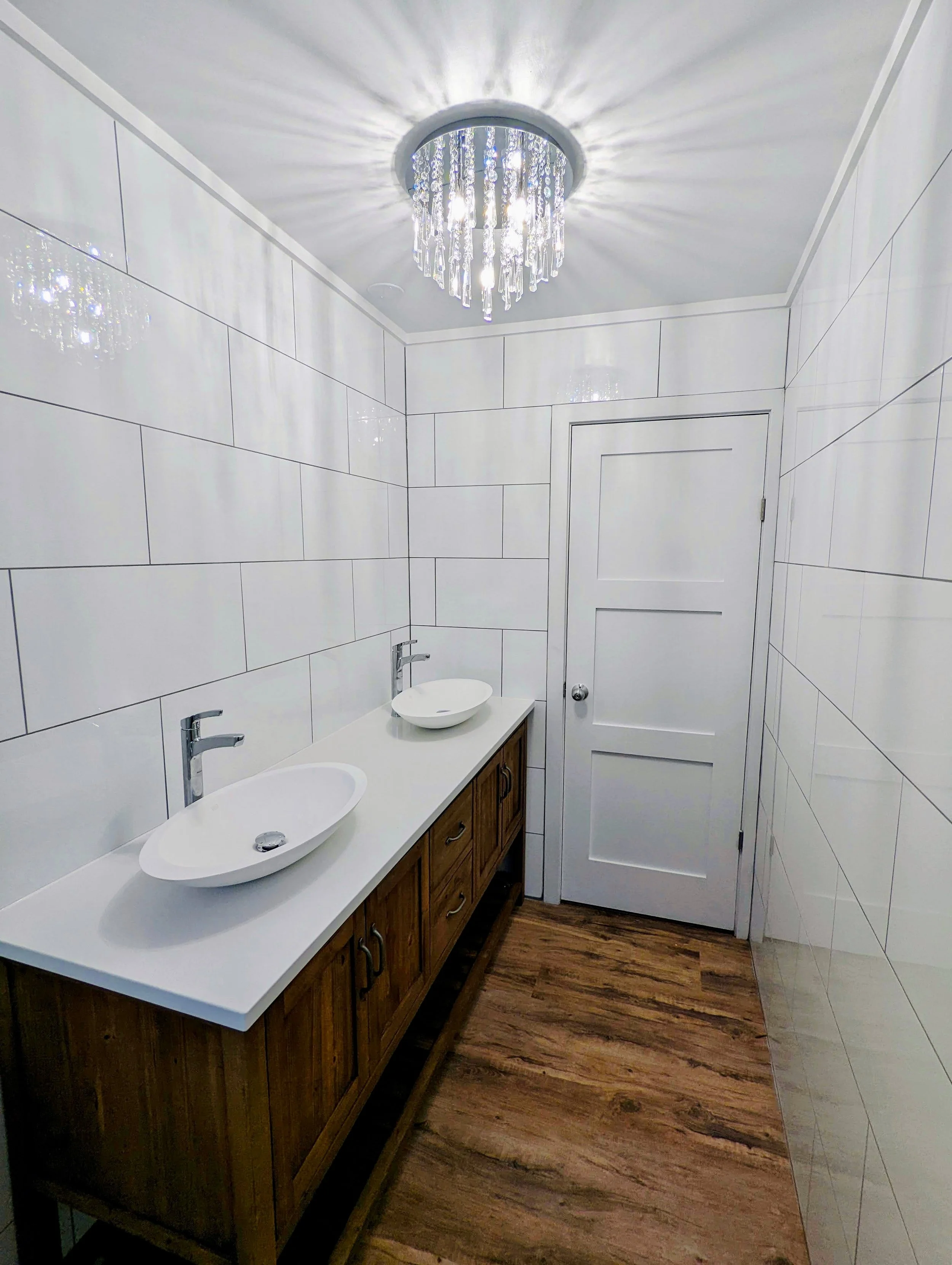 A modern bathroom with a double vanity, round vessel sinks, and a crystal chandelier. The walls are tiled in white, and the wooden floor adds warmth to the space.