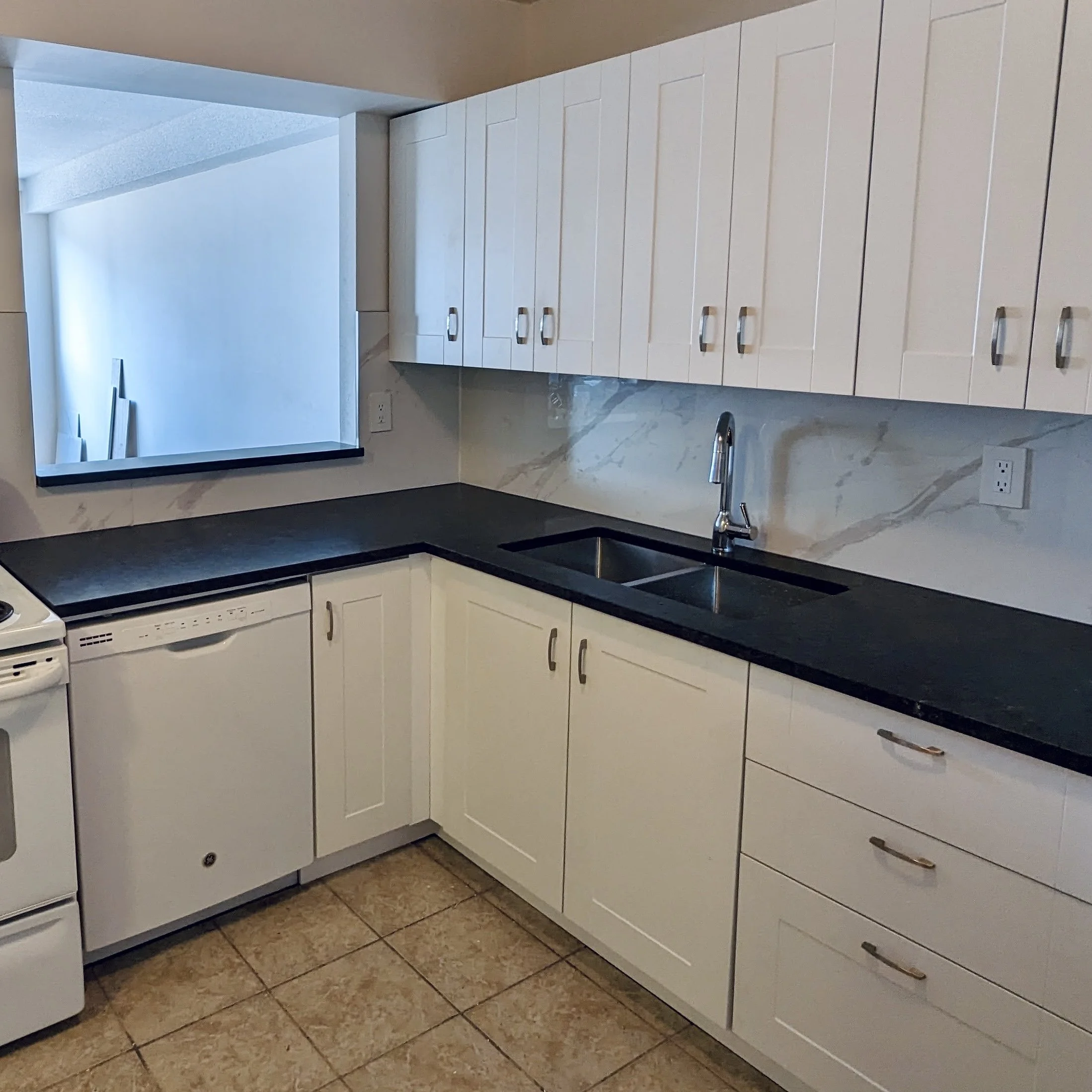 Empty kitchen with white cabinets, black countertop, stainless steel sink, and a window overlooking an interior staircase.